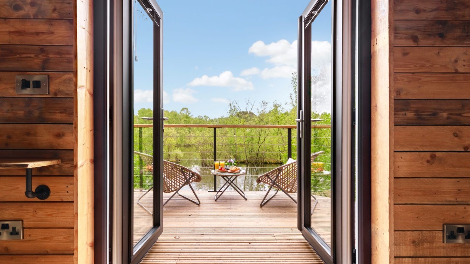 Open double doors lead to a wooden deck with two wicker chairs, offering a Treetop Hideaways view of the lush landscape.