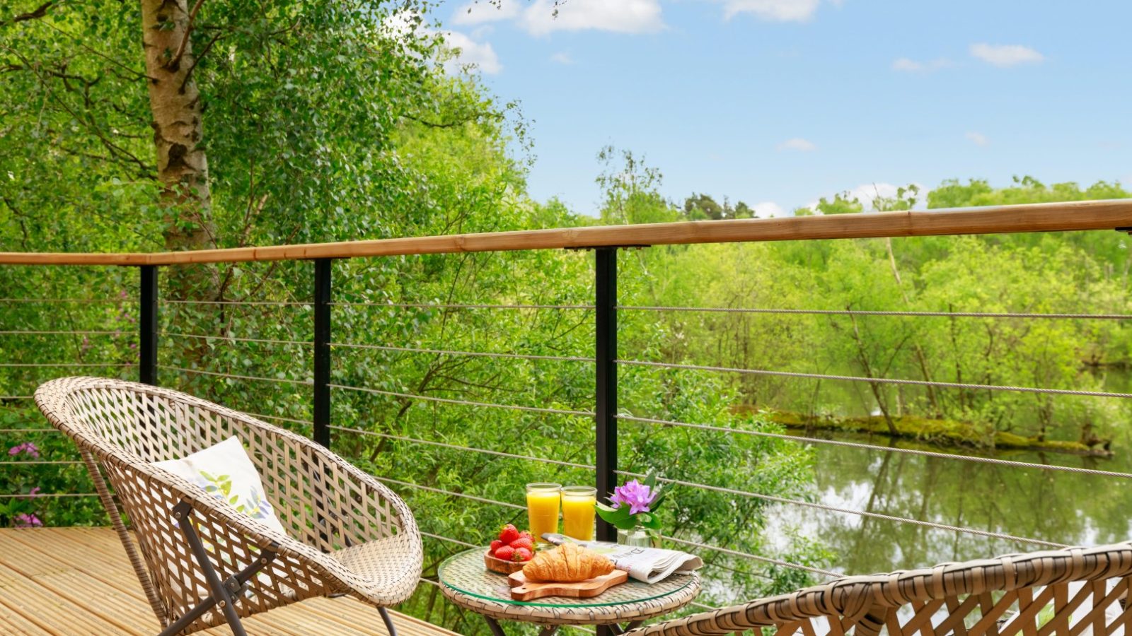 Wicker chairs on a wooden deck at Treetop Hideaways, overlooking forested views. A table holds croissants, strawberries, and orange juice.