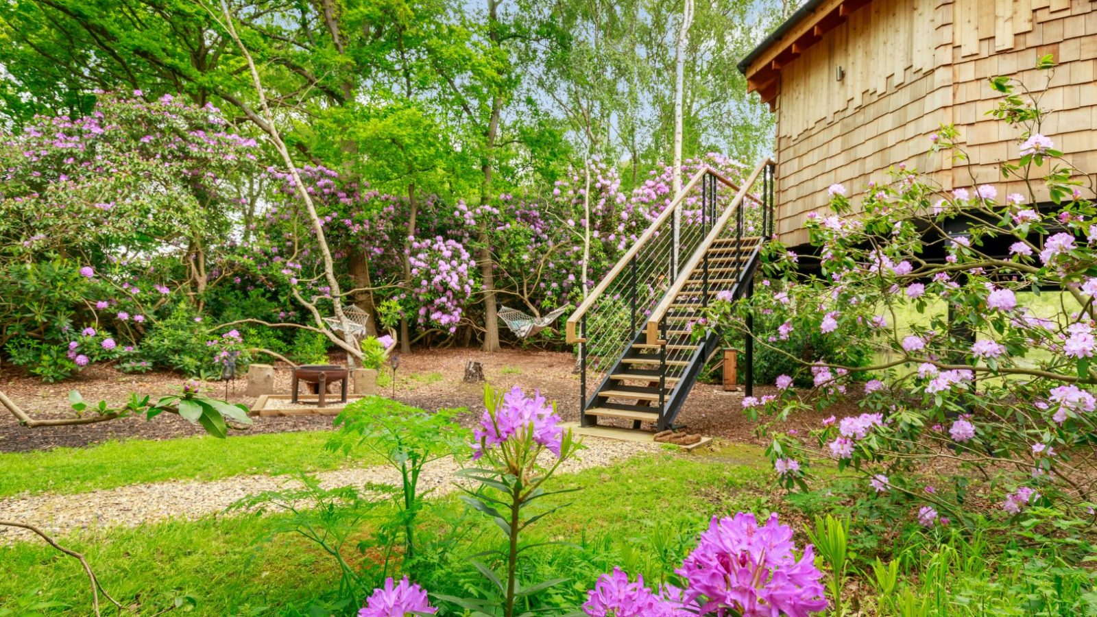 Treetop Hideaways: A wooden treehouse with a staircase amid blooming rhododendrons and green foliage, pathway and benches in view.