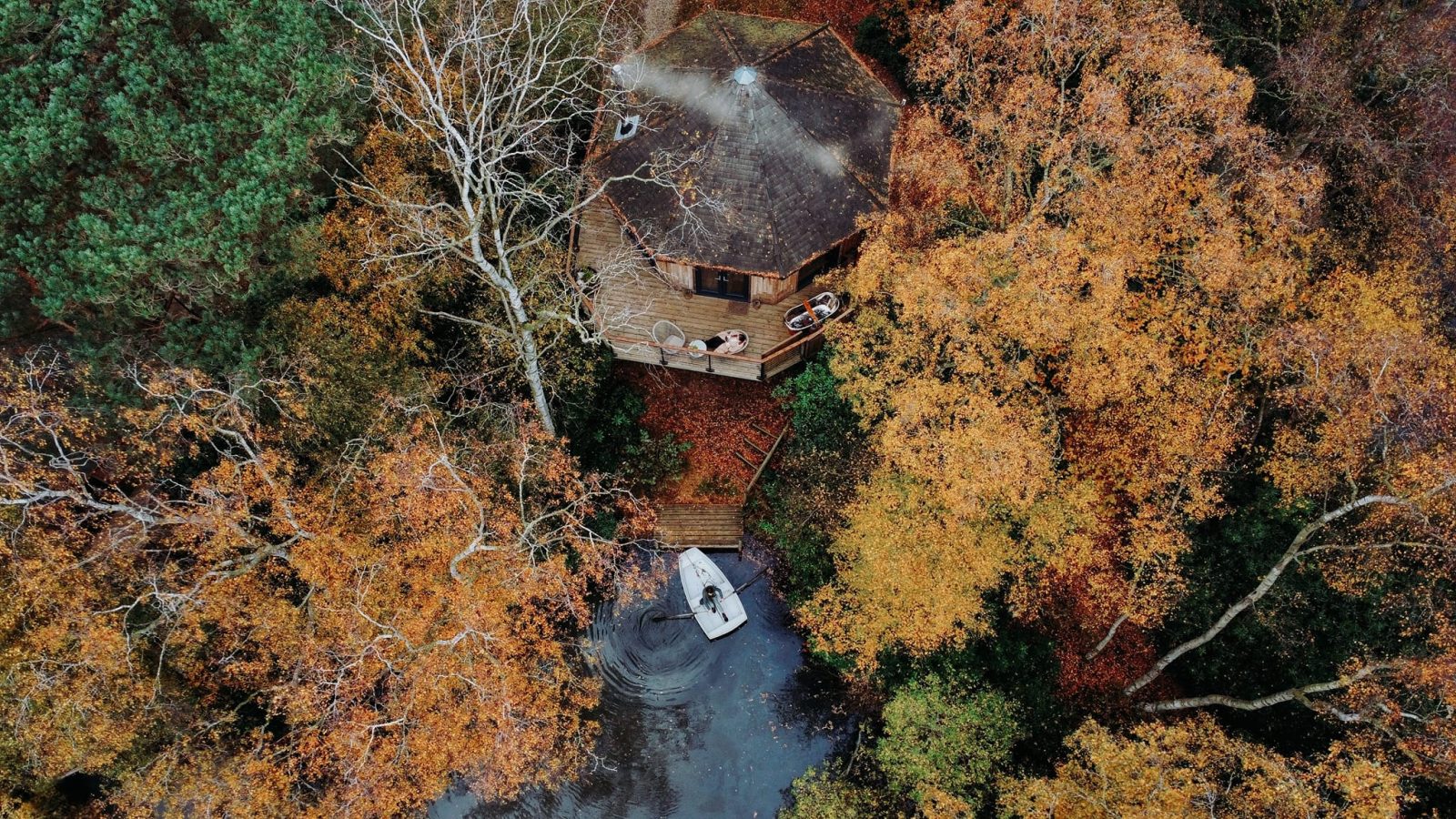 Aerial view of a Treetop Hideaway cabin enveloped by autumn trees, with a small boat on a nearby body of water.