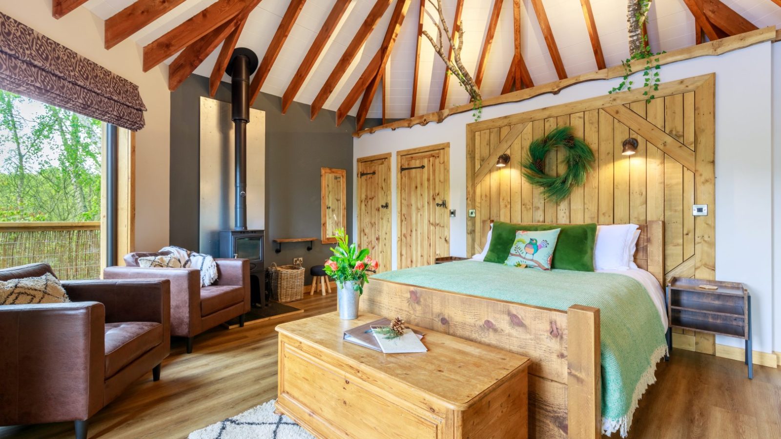 Treetop Hideaways' cozy bedroom has wooden beams, a green-accented bed, armchairs, a wood stove, and a large window showcasing greenery.