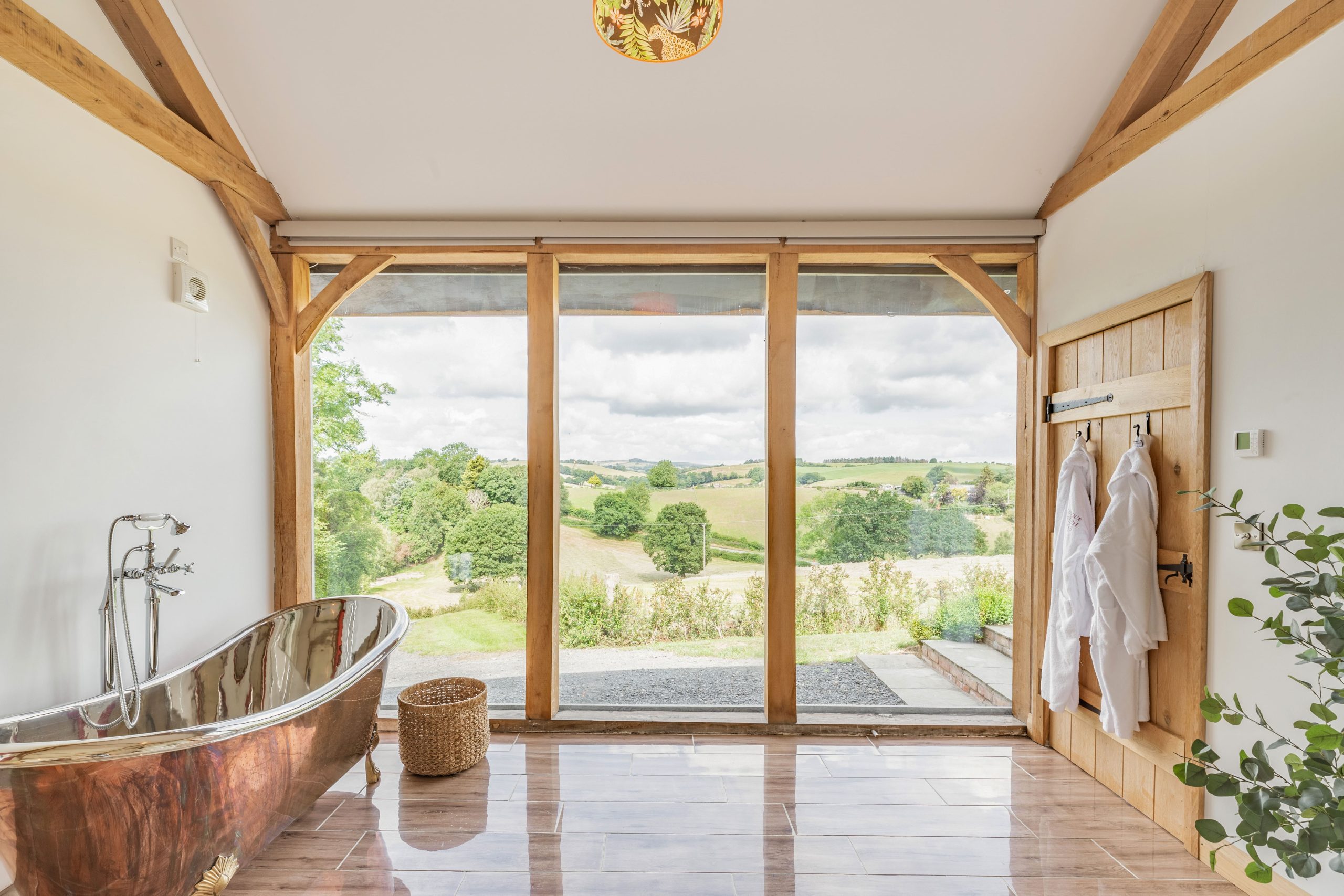 Romantic bathroom with a copper bathtub, wooden beams, and large windows overlooking scenic, dog-friendly landscapes.