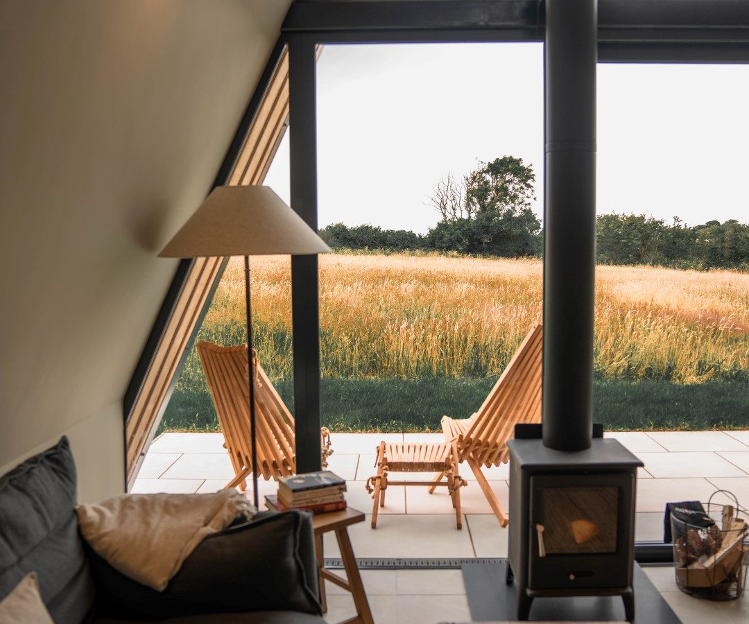 Cozy A-frame cabin interior with a sofa, lamp, and stove. Large windows showcase deck chairs and wilderness views outside.