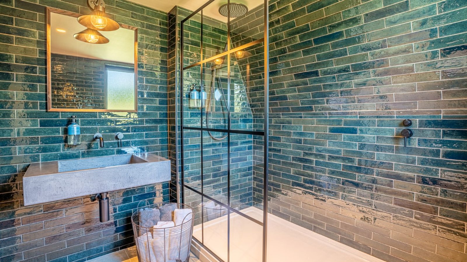 Modern bathroom with green tiled walls, a glass shower, concrete sink, round mirror, and pendant lights—your own nature retreat.