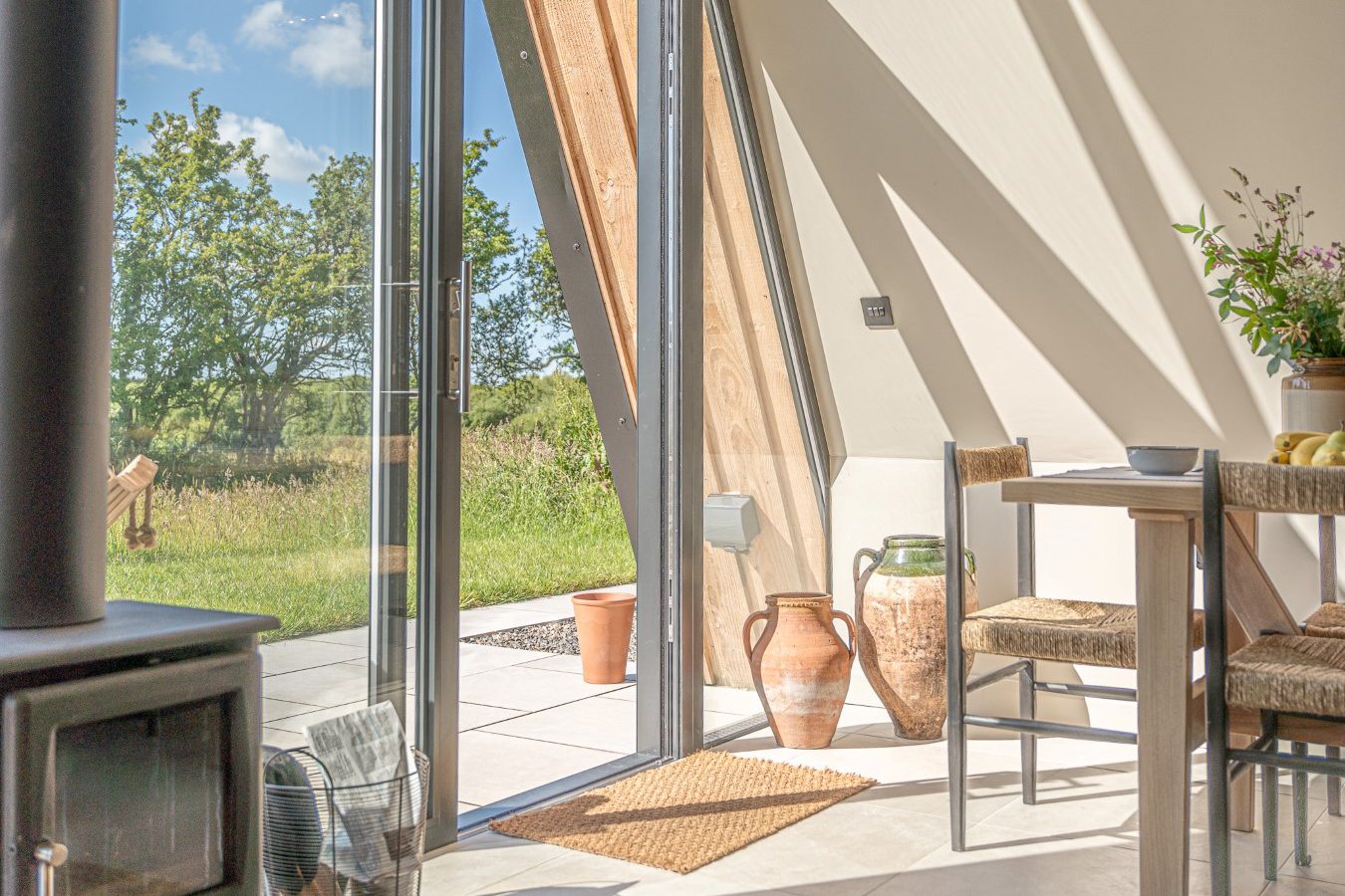 Sunny room with a wood stove at Wilder Retreats; chairs and table near an open glass door leading to a patio garden view.