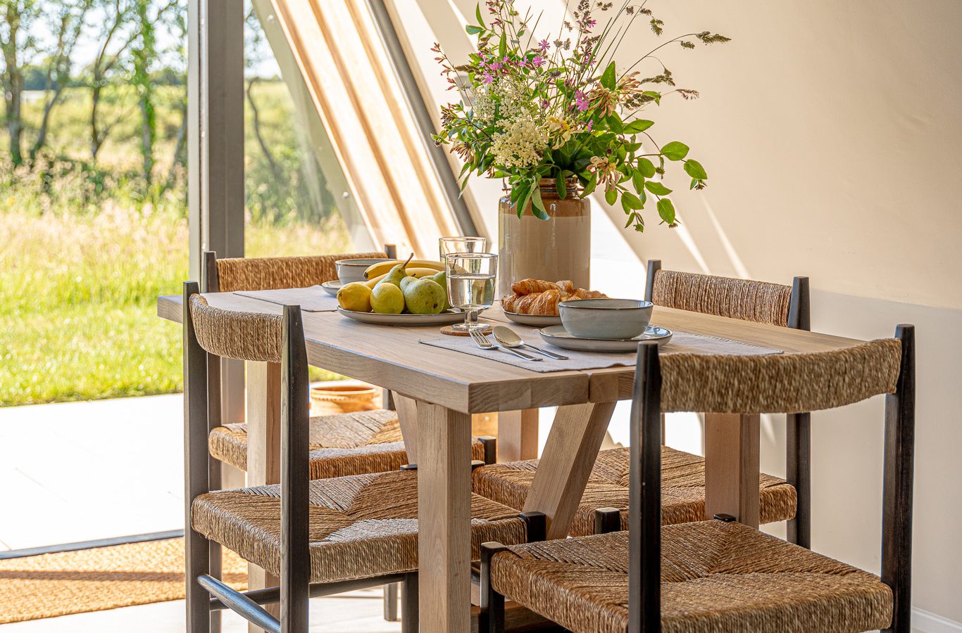 A wooden dining table by a large window, adorned with flowers, apples, and pastry—perfect for a cozy Wilder Retreats escape.