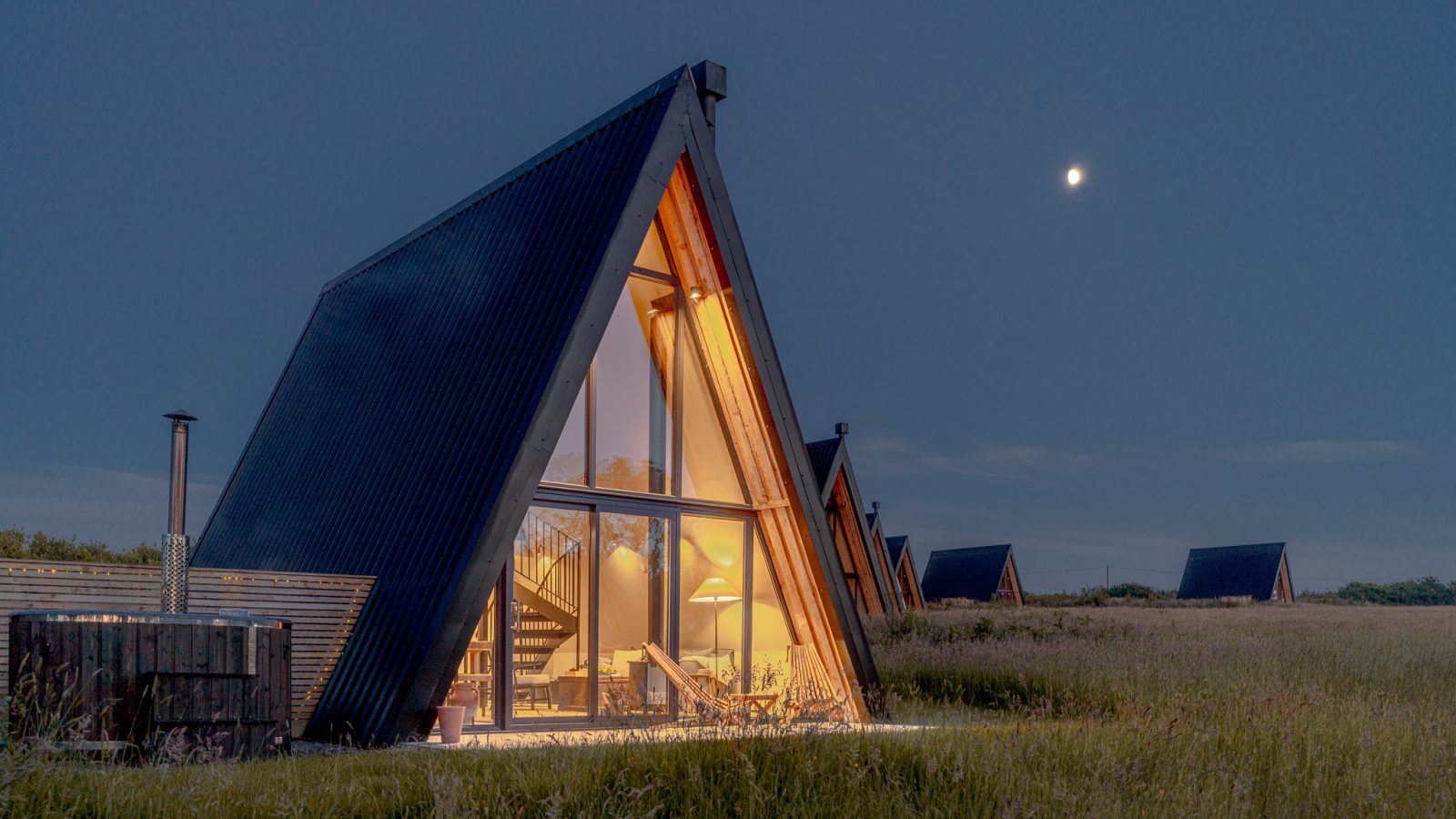 A-frame cabin with illuminated interior at dusk, set in a grassy field under a clear night sky at Wilder Retreats.