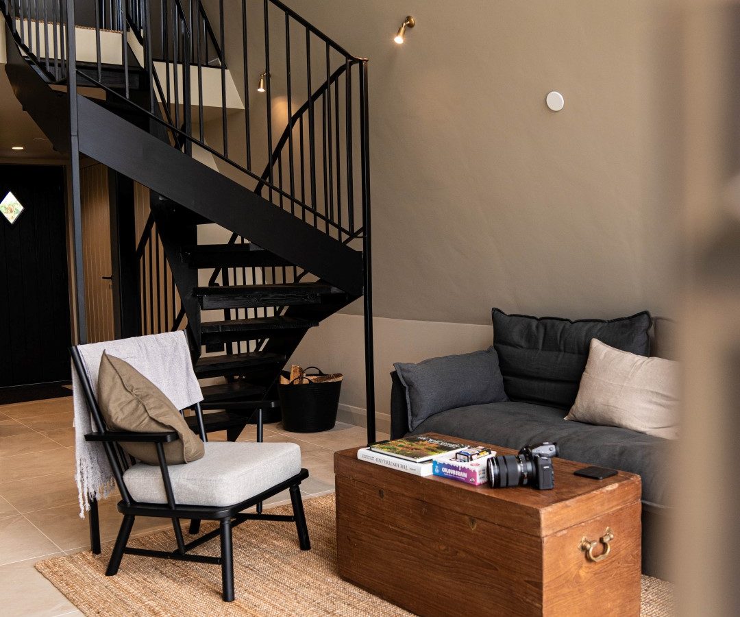 Living room at Wilder Retreats: spiral staircase, gray couch, wooden chest, and chair on a woven rug. Neutral walls and tiled floor.