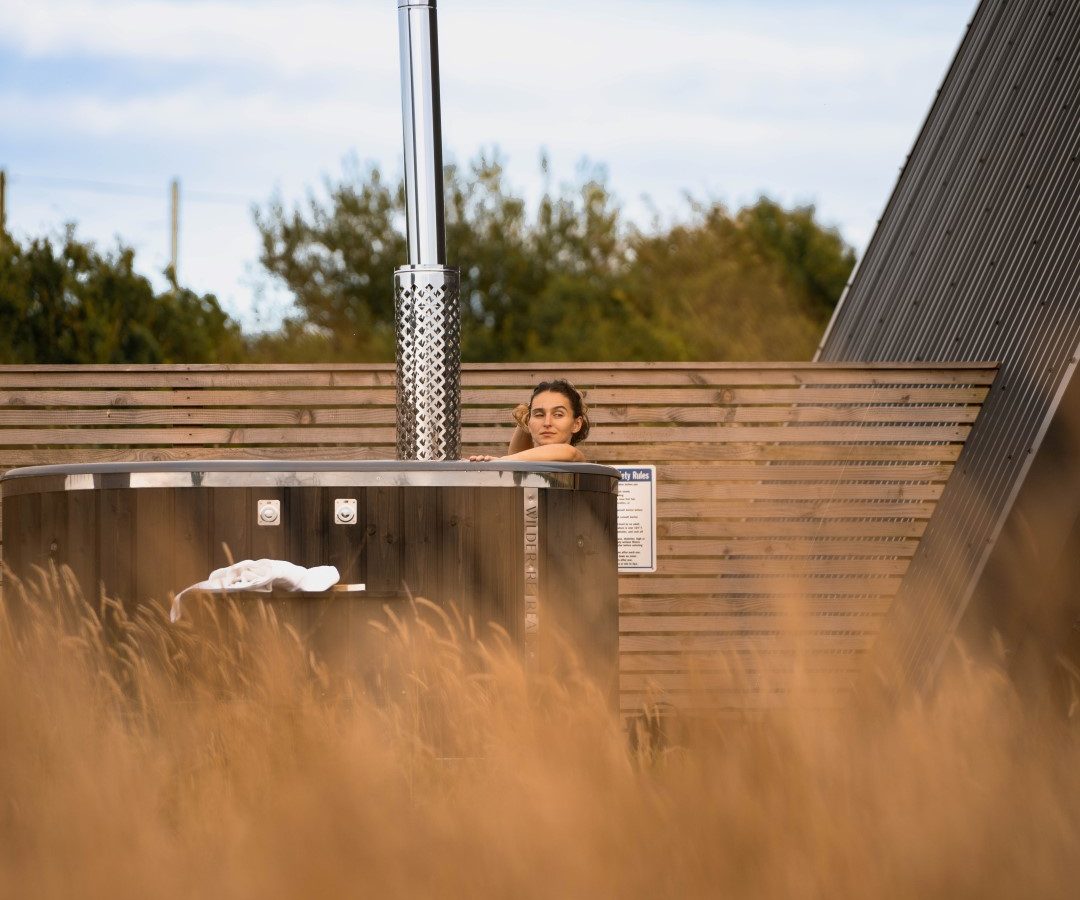 Person in a wooden hot tub outside, obscured by grasses, enjoying a Wilder Retreats escape with a chimney against a clear sky.