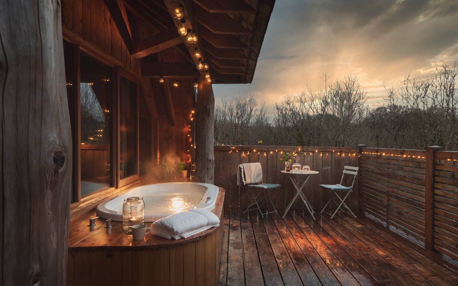 Romantic outdoor deck with a hot tub, small table, chairs, string lights, and trees in the sunset—ideal for dog-friendly breaks.