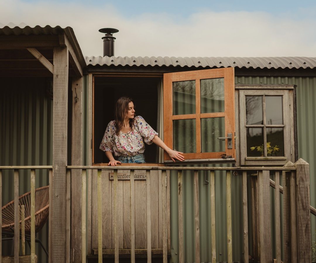 A woman stands on a wooden porch in Tokenhill, Dorset, opening a half-door against corrugated metal walls and a wooden railing.