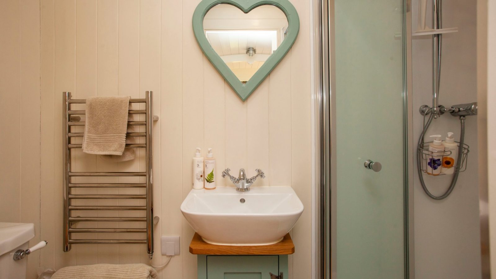 Bathroom in Tokenhill Dorset with a sink, green heart-shaped mirror, towel rack, shower stall, and toiletries on a wooden cabinet.