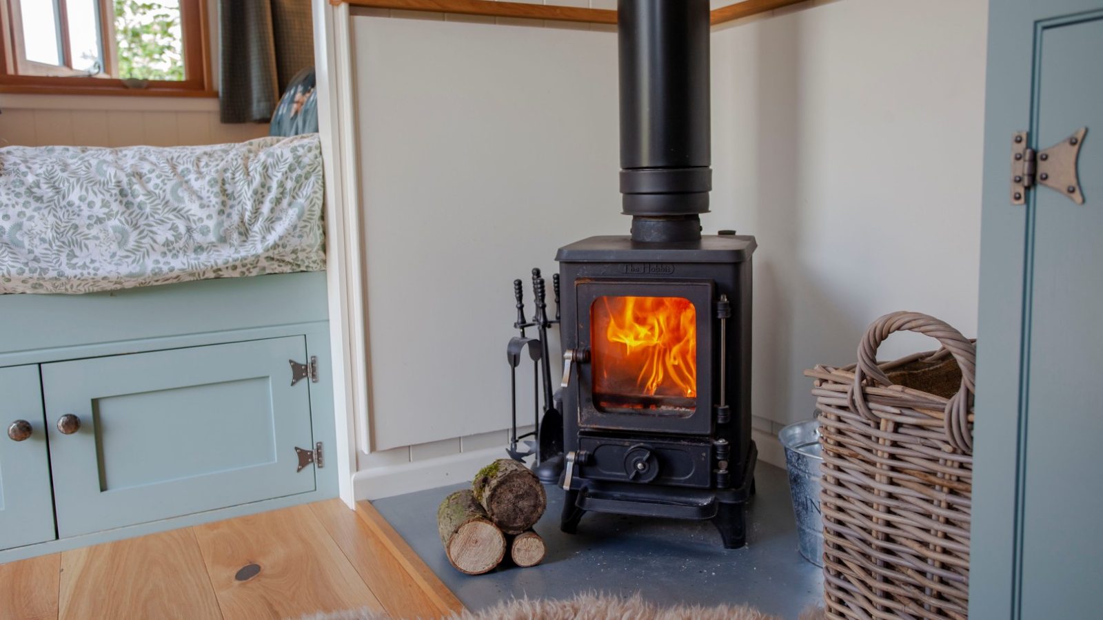 Cozy room in Tokenhill Dorset: wood-burning stove, visible flames, logs, woven basket, and a bed with green cabinets.