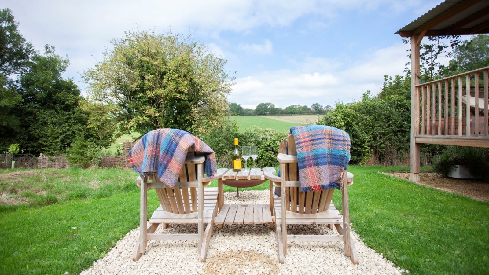 Two wooden chairs with plaid blankets at Tokenhill, Dorset face a green landscape; a table holds a bottle and two glasses.