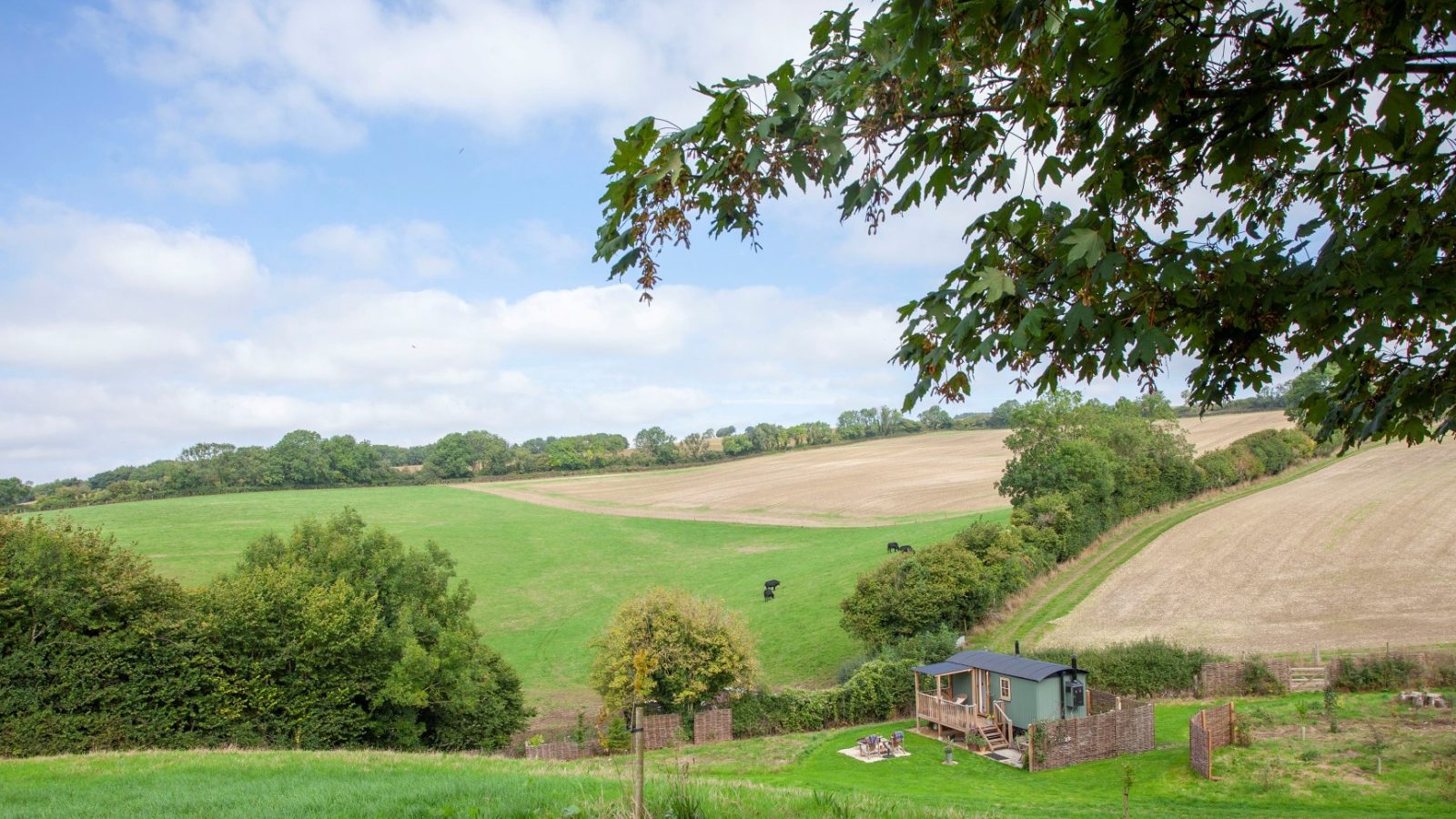 Countryside scene in Tokenhill, Dorset with rolling fields, a small cabin, trees, and a partly cloudy sky.