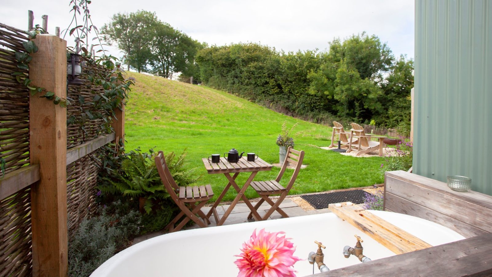 Outdoor setup with a Tokenhill Dorset vibe: wooden table and chairs on a patio, wooden bathtub with pink flower, green lawn, trees.