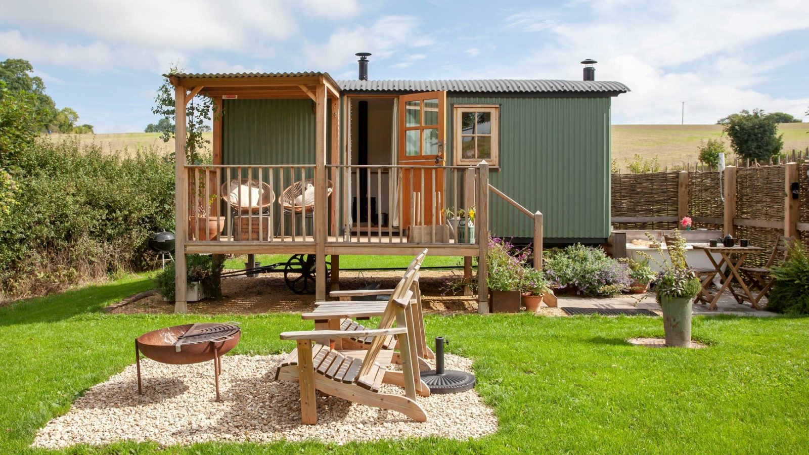 A green shepherd's hut in Tokenhill, Dorset, with a porch and chairs, nestled in a garden with a nearby fire pit.