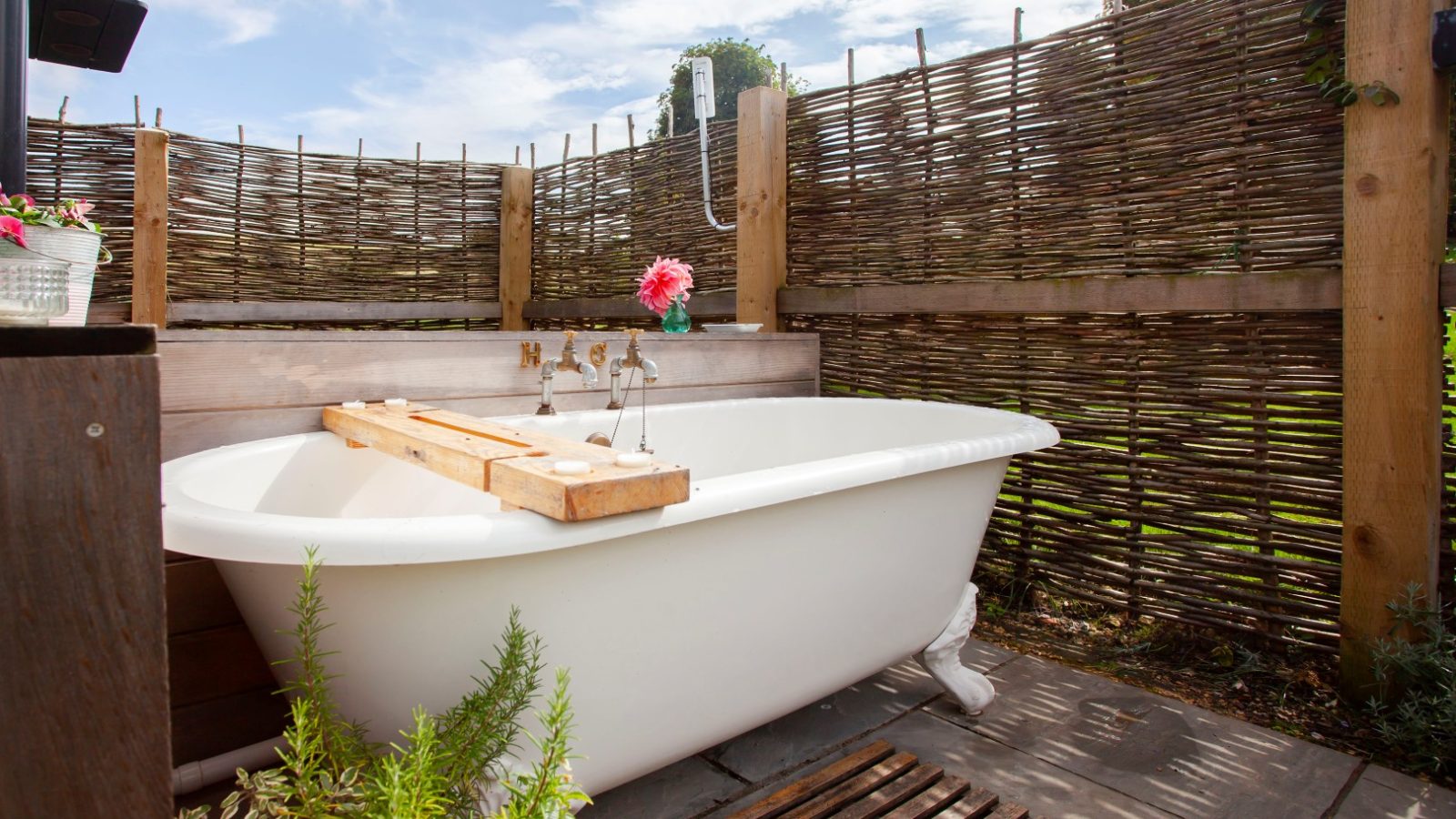 Outdoor clawfoot bathtub in rustic Tokenhill, Dorset, with wicker fencing and wooden tray, surrounded by plants under a blue sky.