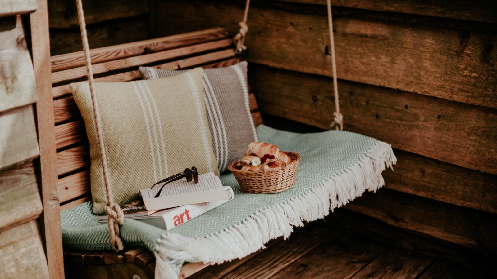 Wooden swing with cushions, glasses, and a basket of pastries on a teal fringed blanket.