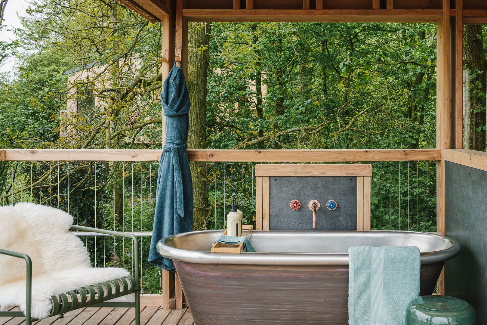 Dog-friendly glamping breaks with an outdoor bathtub on a wooden deck, blue towel, green chair, and lush greenery.