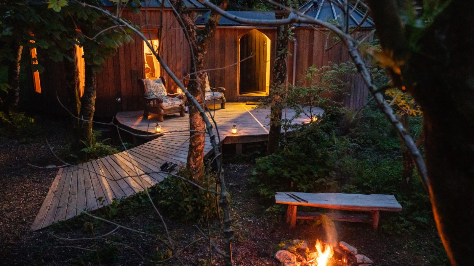 A wooden cabin, nestled in the forest with pathways, a fire pit, and candles illuminating the deck at dusk. Little Menherion Glamping Cornwall