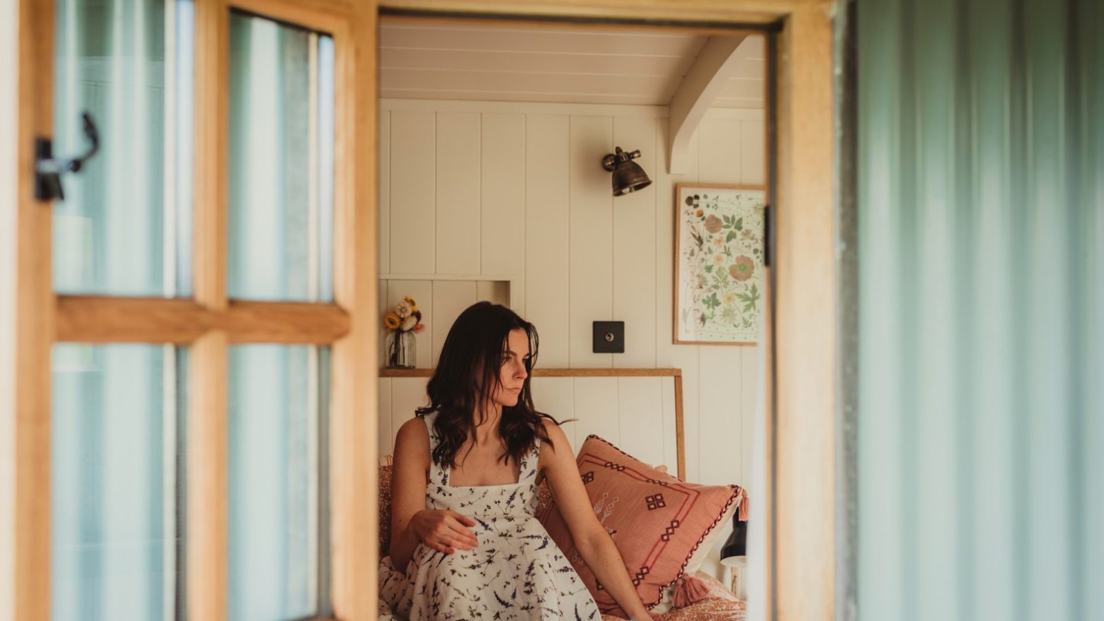 In Tokenhill Dorset, a woman in a floral dress sits on a bed, seen through an open window in a cozy room with light walls.