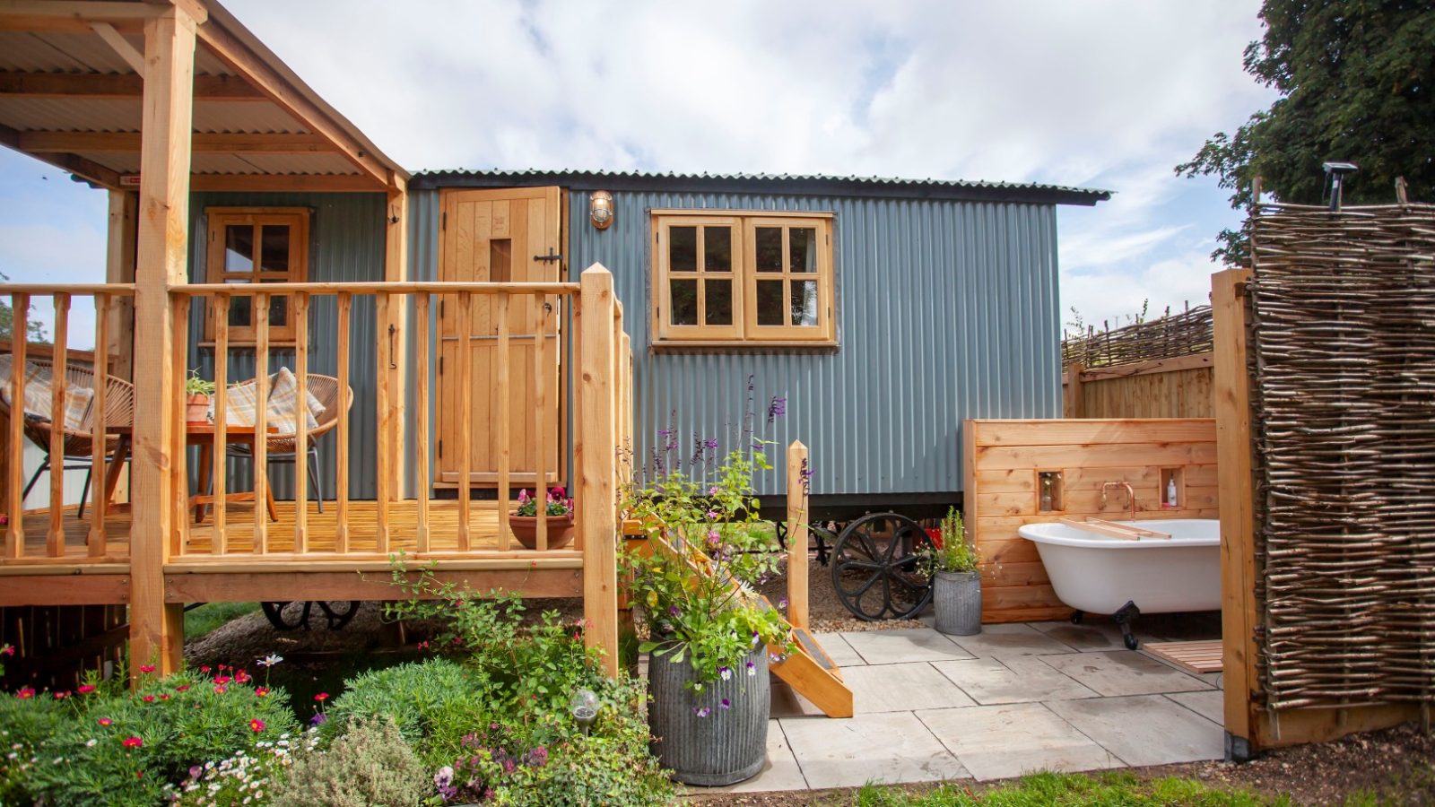 A small blue corrugated metal cabin in Tokenhill, Dorset, with a wooden deck and an outdoor bathtub on stone tiles.