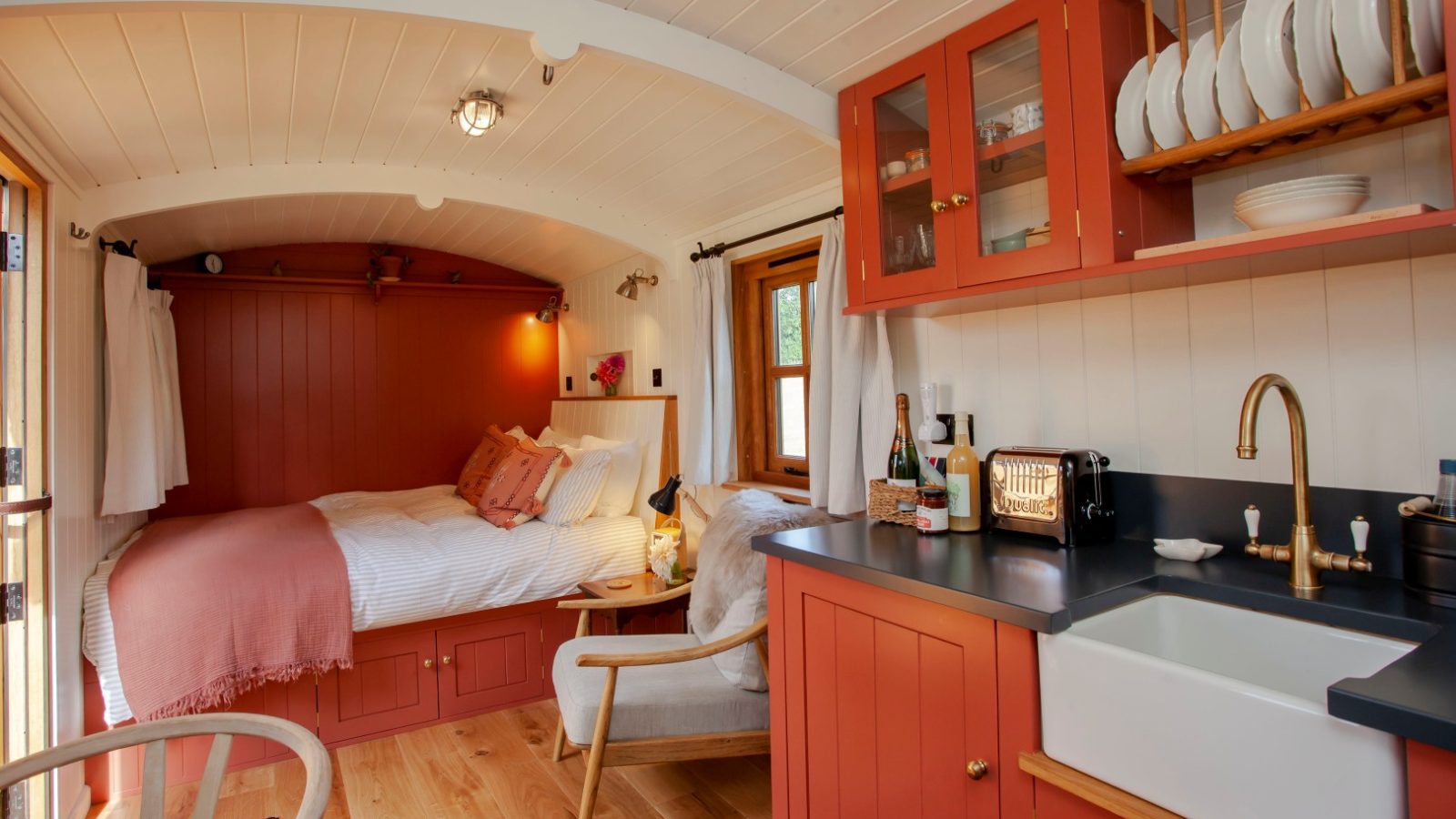 Compact and cosy tiny home interior in Tokenhill Dorset, with a red and white theme, featuring a bed, kitchenette, and wooden chair.