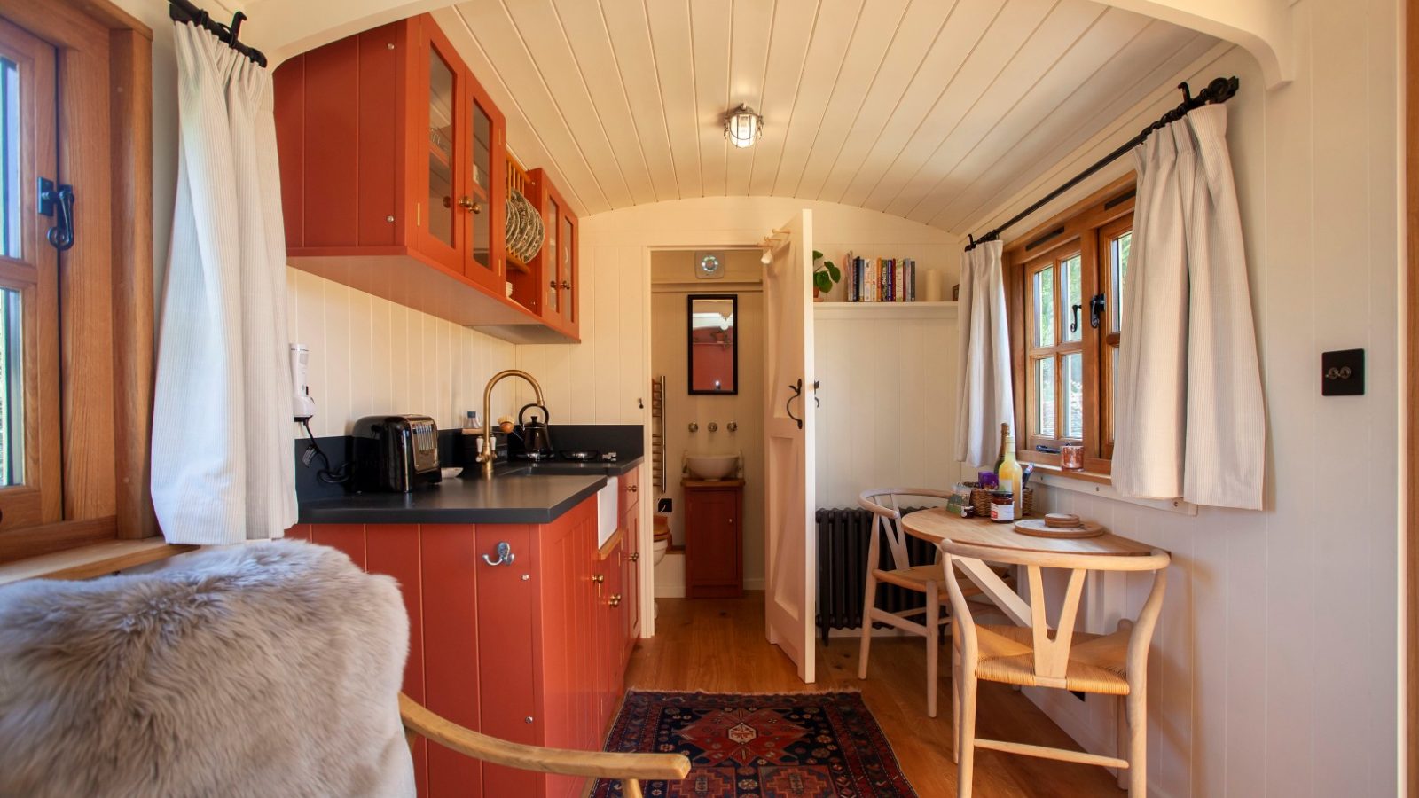 Cozy Tokenhill Dorset cabin interior with red cabinets, wooden chairs, a rug, and windows framed by white curtains.