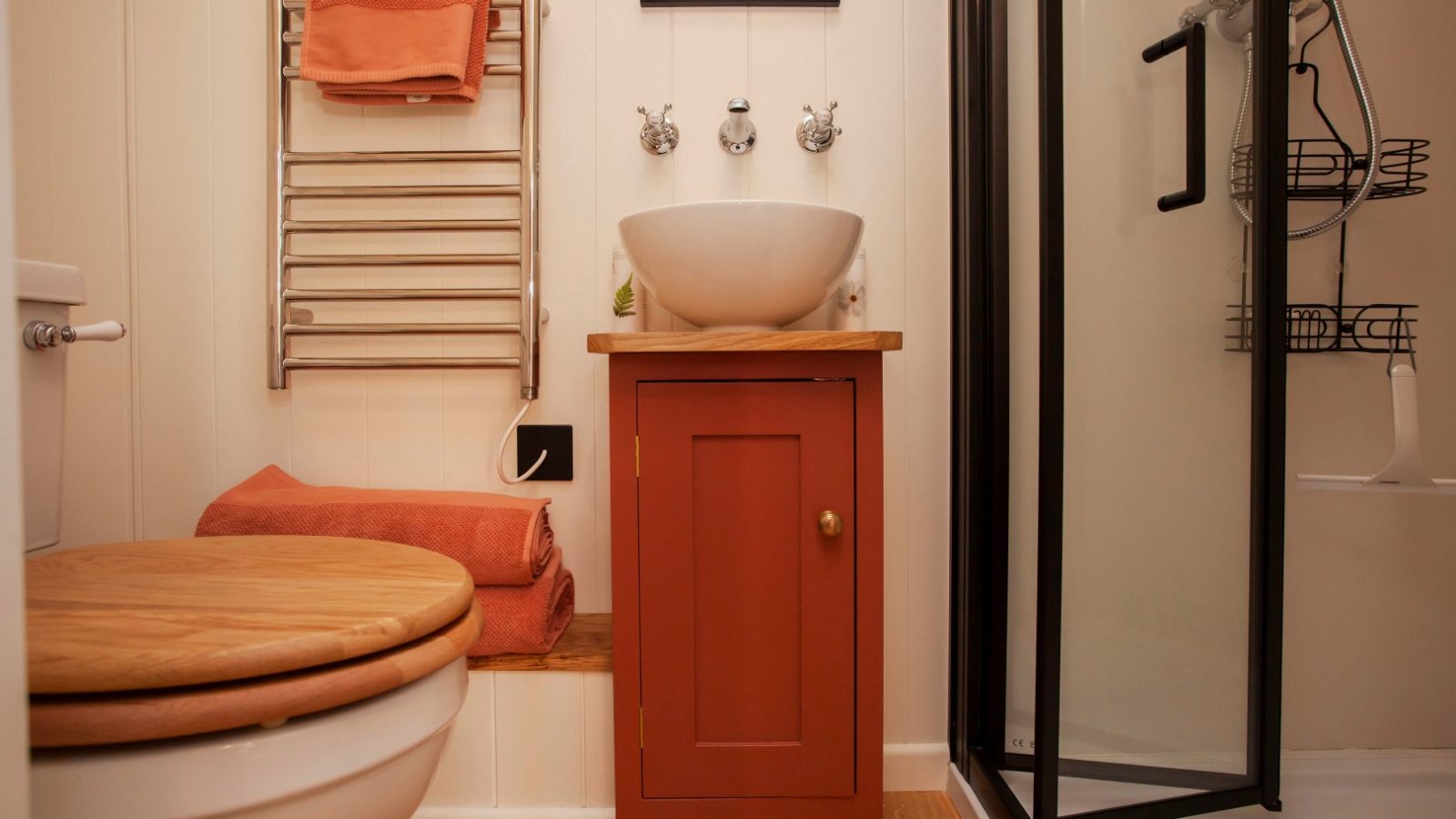 Compact Tokenhill Dorset bathroom with a wooden toilet seat, red cabinet, round sink, wall mirror, and glass shower enclosure.