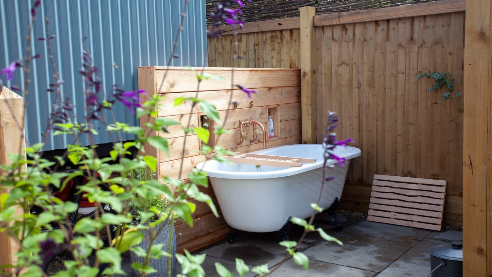 Outdoor bathtub with wooden privacy walls, nestled in Tokenhill Dorset's greenery and flowers on a stone-tiled area.