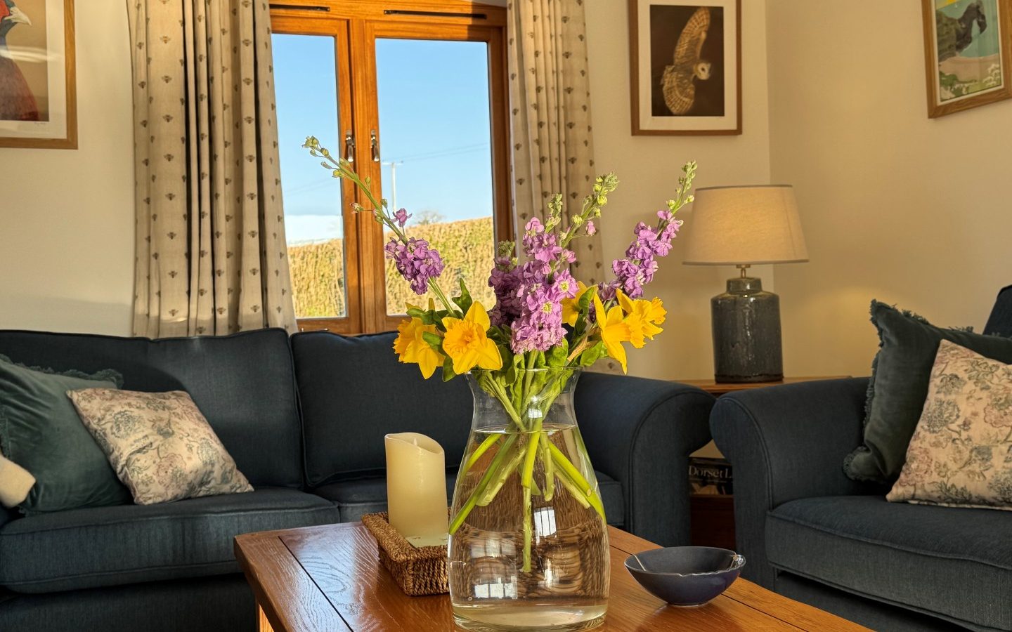 In a well-lit Tokenhill Dorset living room, yellow and purple flowers grace a wooden table surrounded by blue sofas.