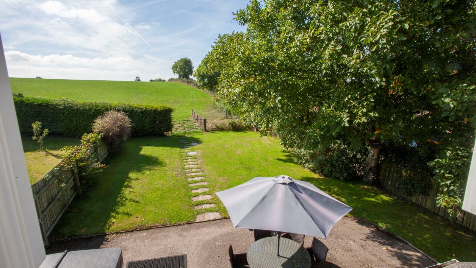 Aerial view of a Tokenhill Dorset backyard with patio table, umbrella, tree, hedge, lawn, and stone path in a rural setting.