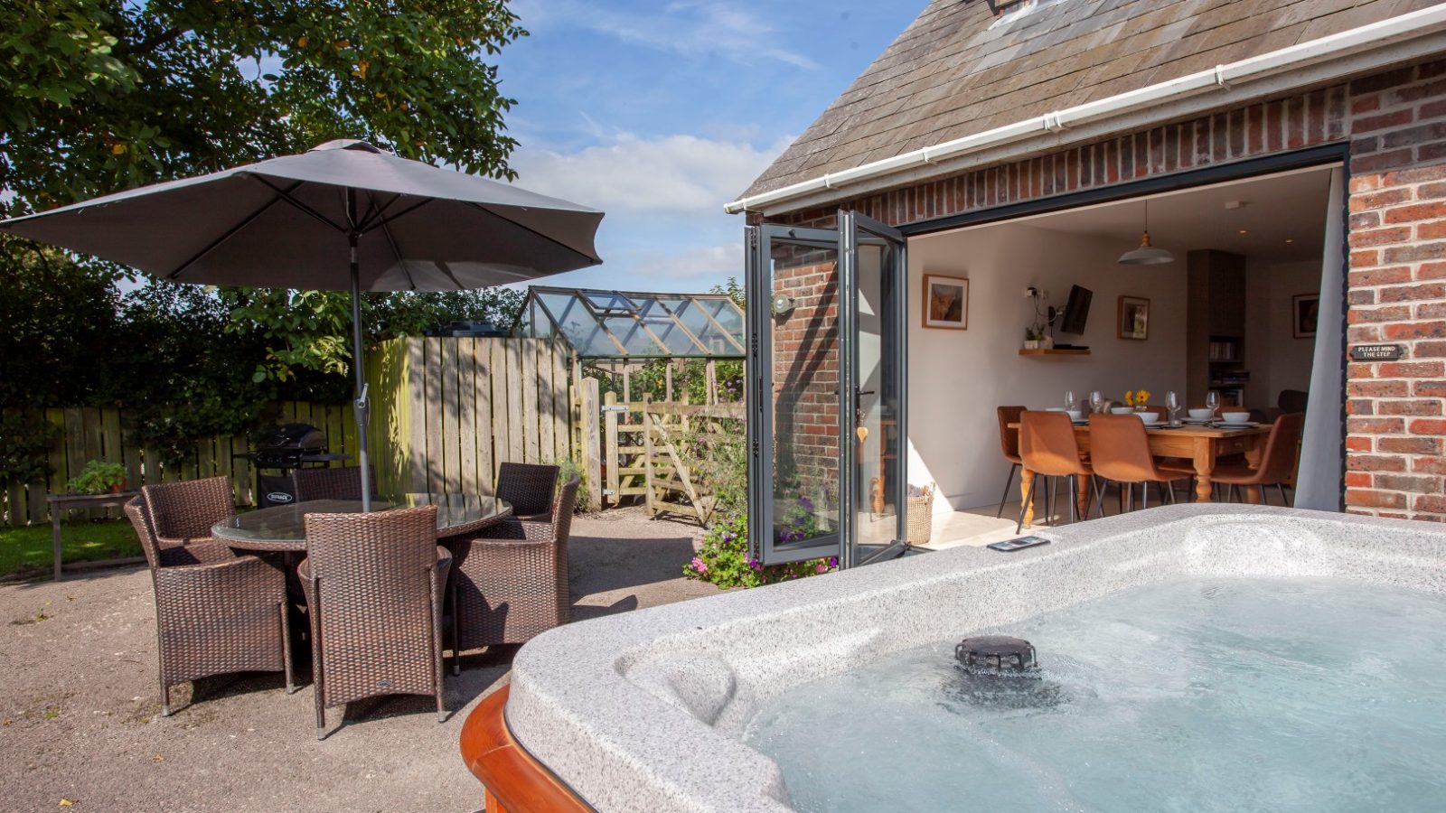 Tokenhill Dorset backyard with hot tub, outdoor dining set under umbrella, greenhouse, and open doors to indoor dining.