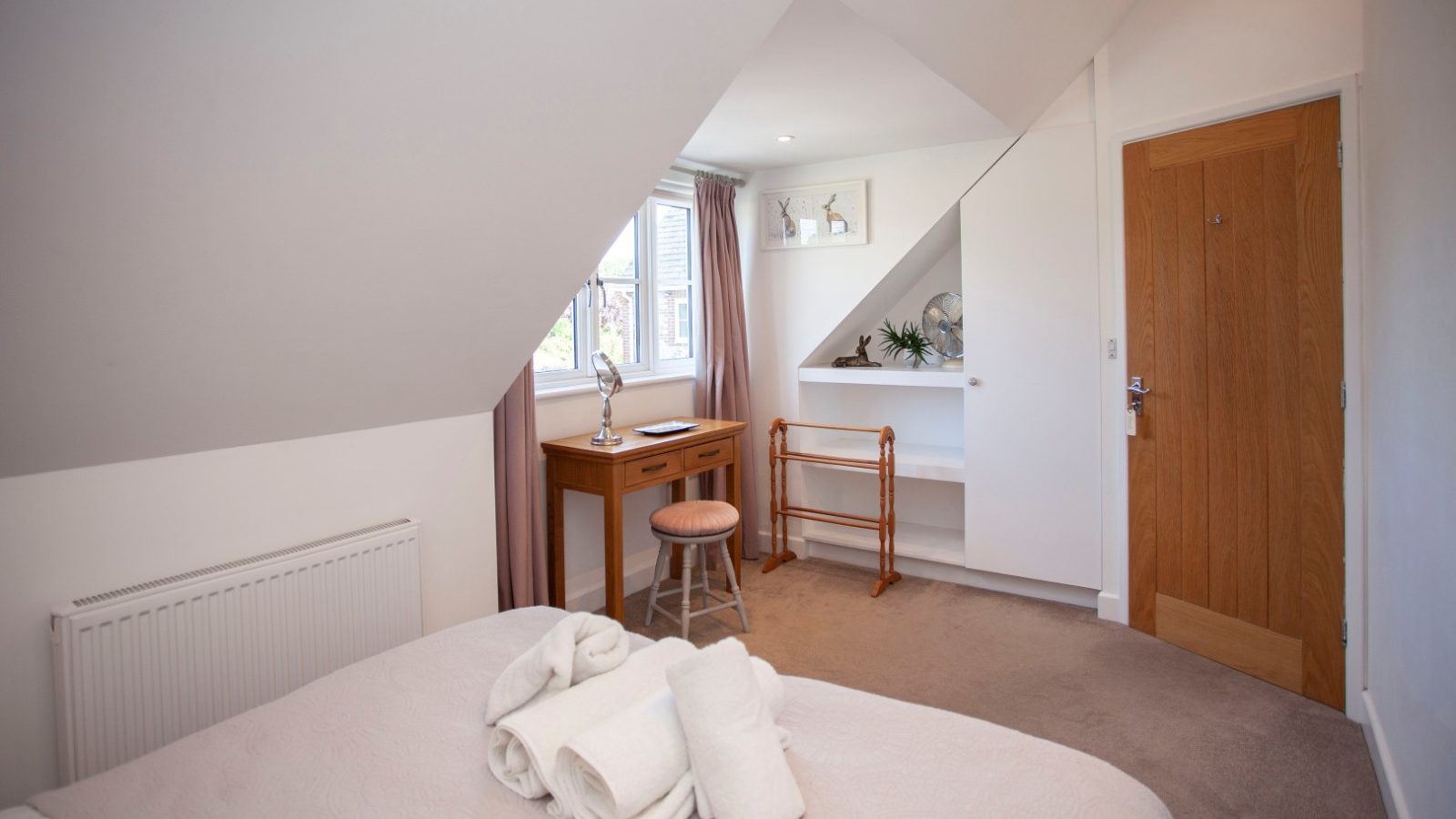 Attic bedroom in Tokenhill, Dorset, with sloped ceiling, double bed, vanity desk, stool, and open wooden door; towels on bed.