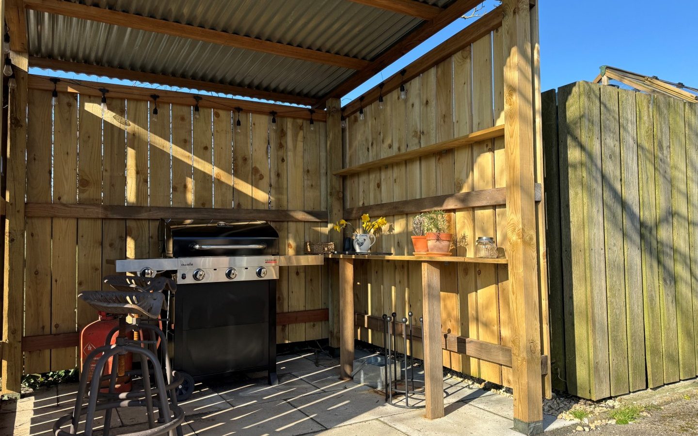 Outdoor wooden barbecue shelter with a grill, stools, and potted plants in sunny Tokenhill, Dorset.