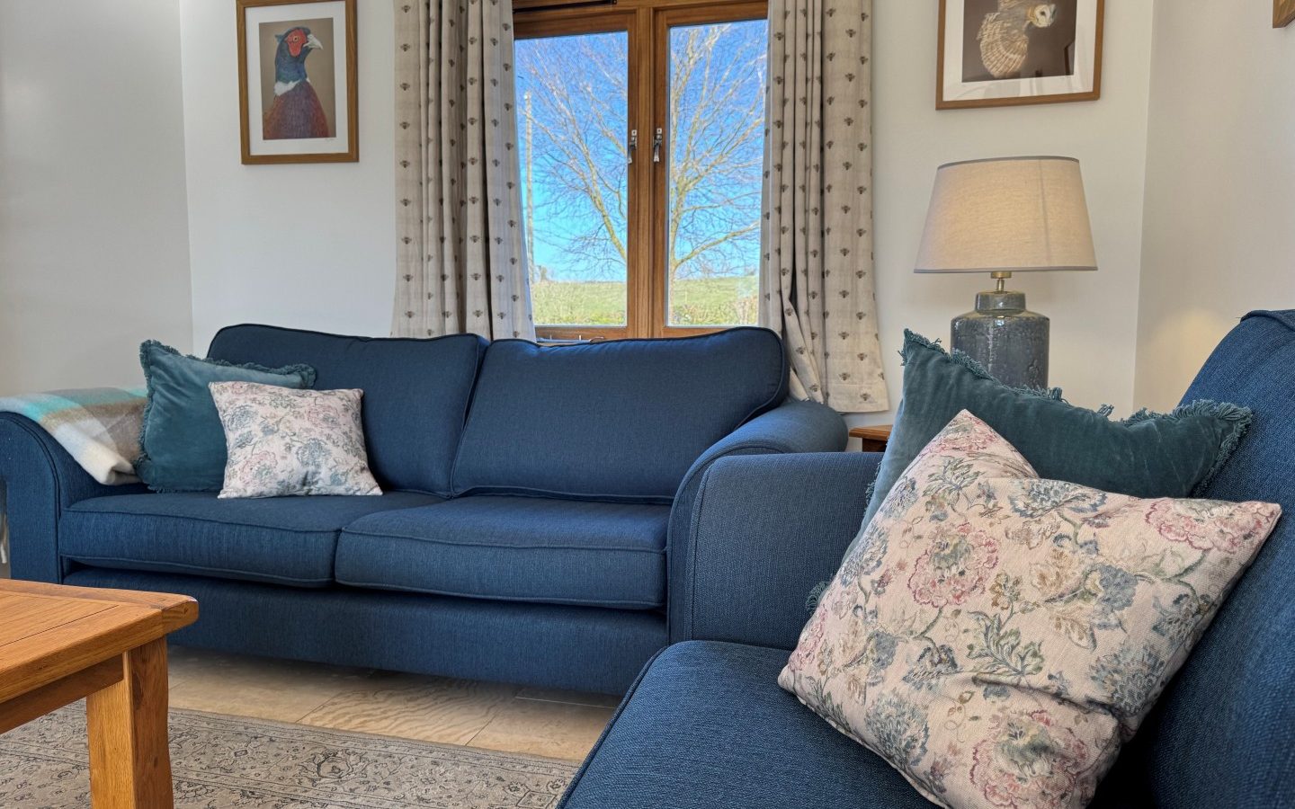 A Tokenhill Dorset living room with blue sofas, floral cushions, a wooden coffee table, and bird artwork near patterned curtains.