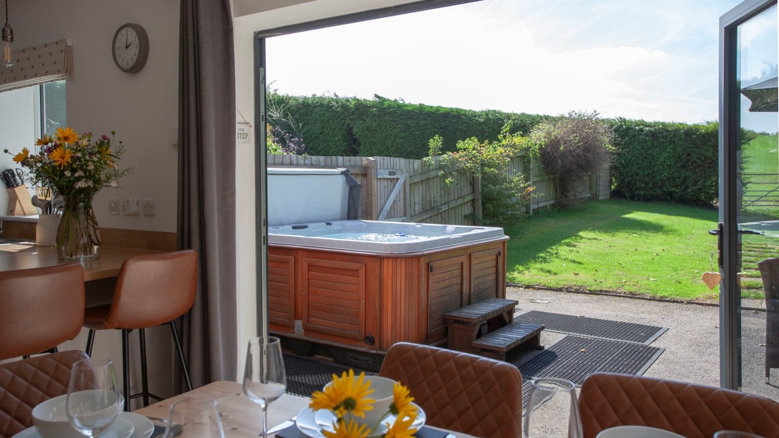 Dining area with a set table and sunflowers. Open patio doors reveal a hot tub, garden, and Tokenhill Dorset's lush green lawn.