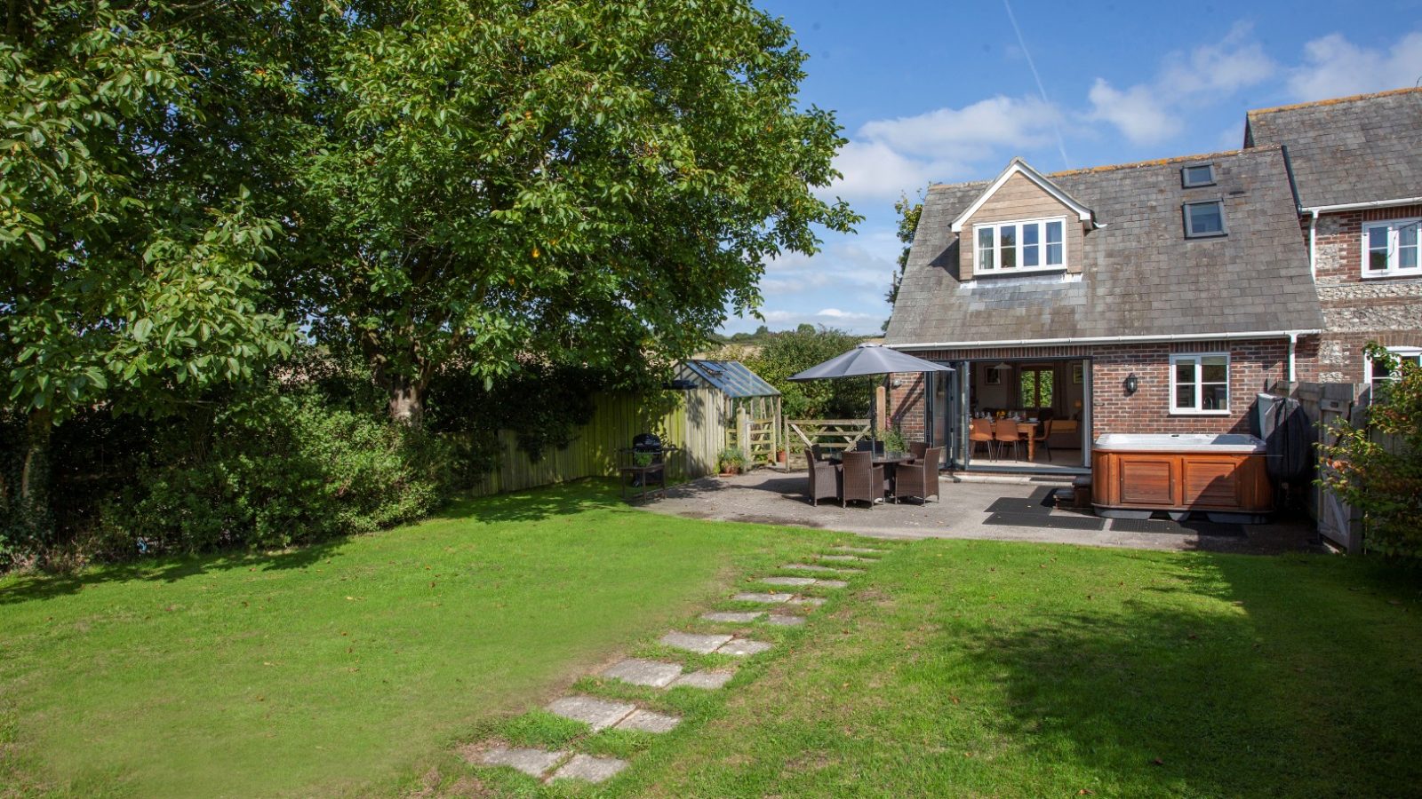 Nestled in Tokenhill Dorset, a backyard scene unfolds with a lawn, patio table, hot tub, and brick house under clear blue sky.