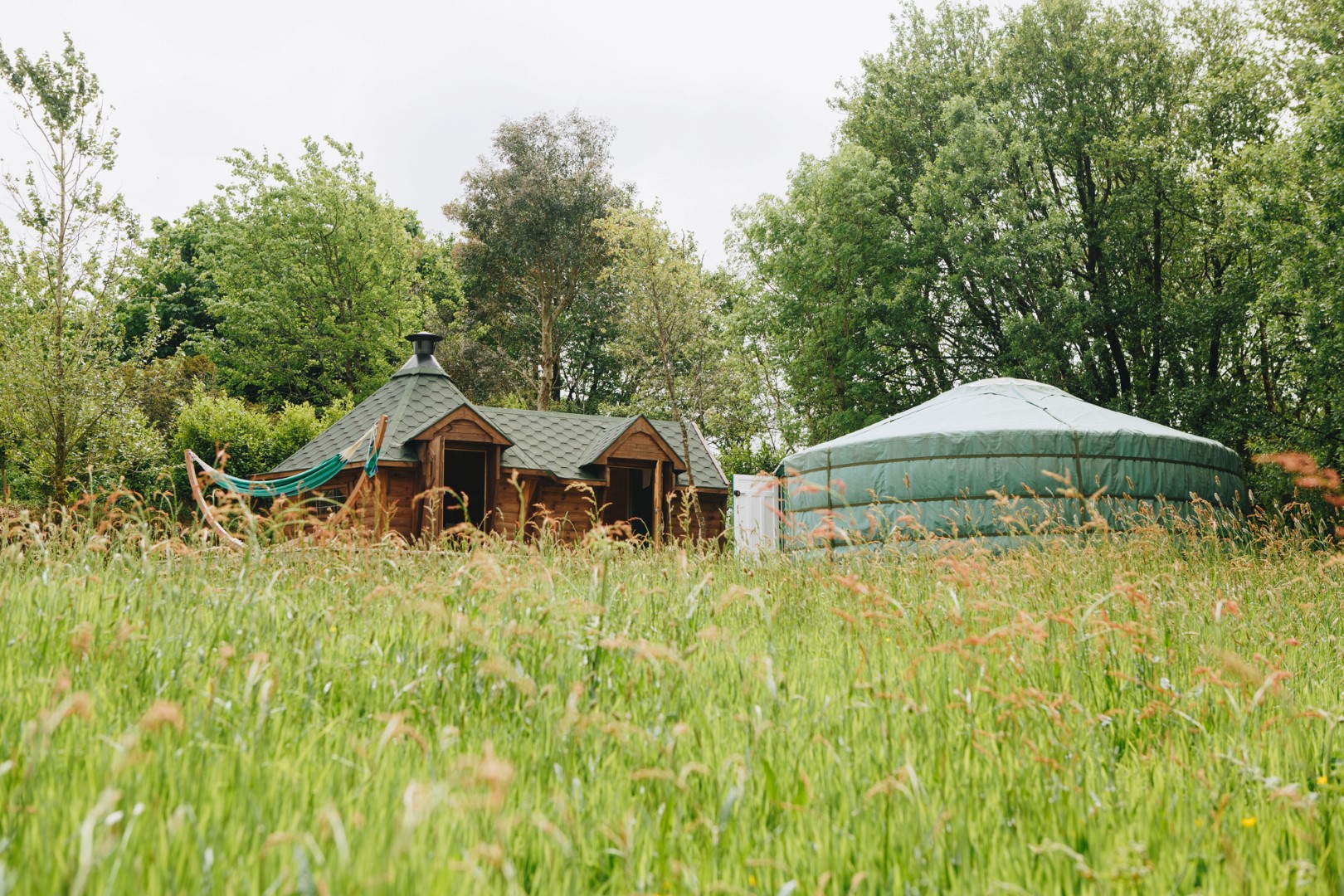 A small wooden cabin and a yurt stand in a wildflower meadow, offering the perfect UK getaway under a cloudy sky.