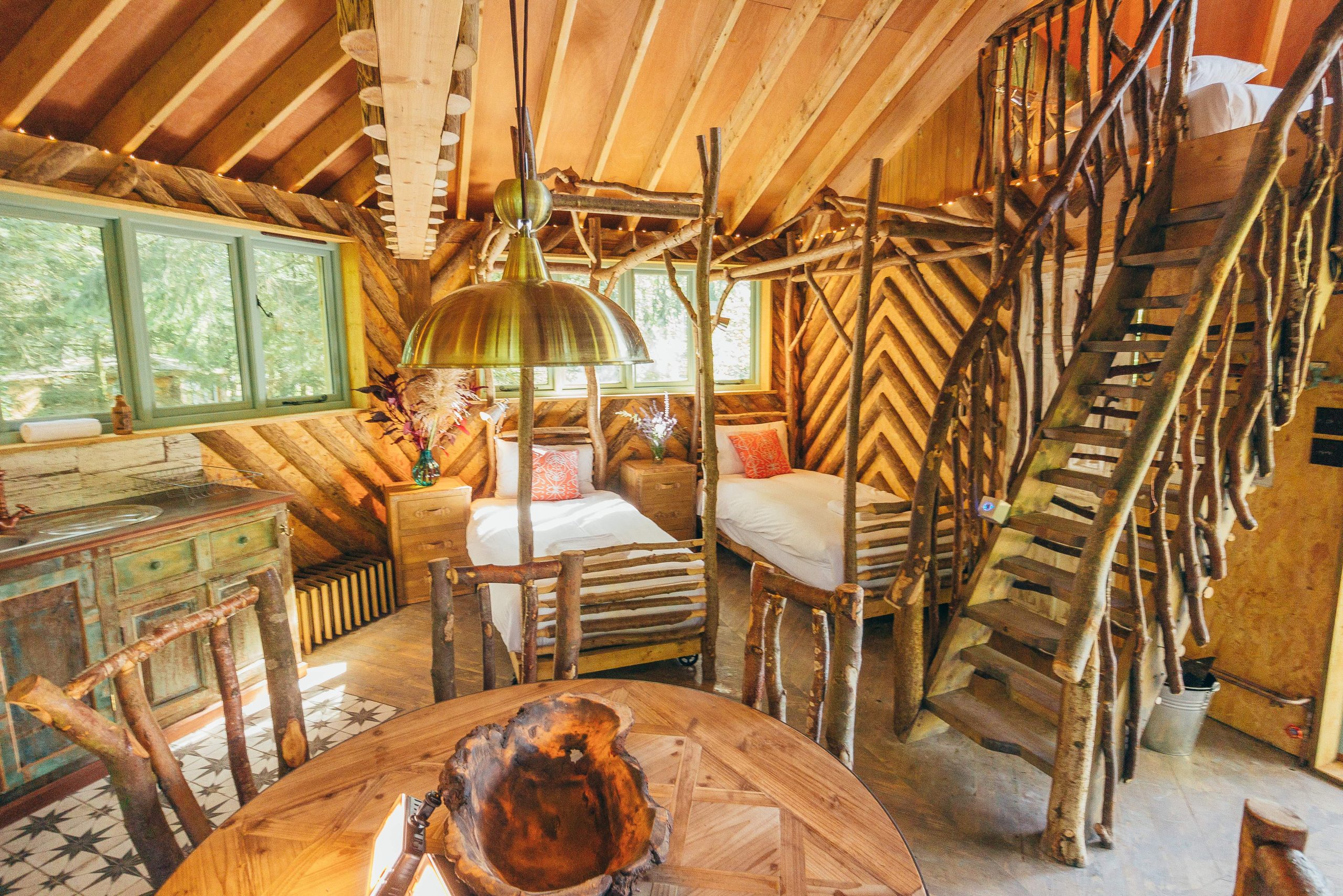 Rustic, dog-friendly cabin interior with twin beds, wooden furniture, spiral staircase, dining table, and large pendant light.