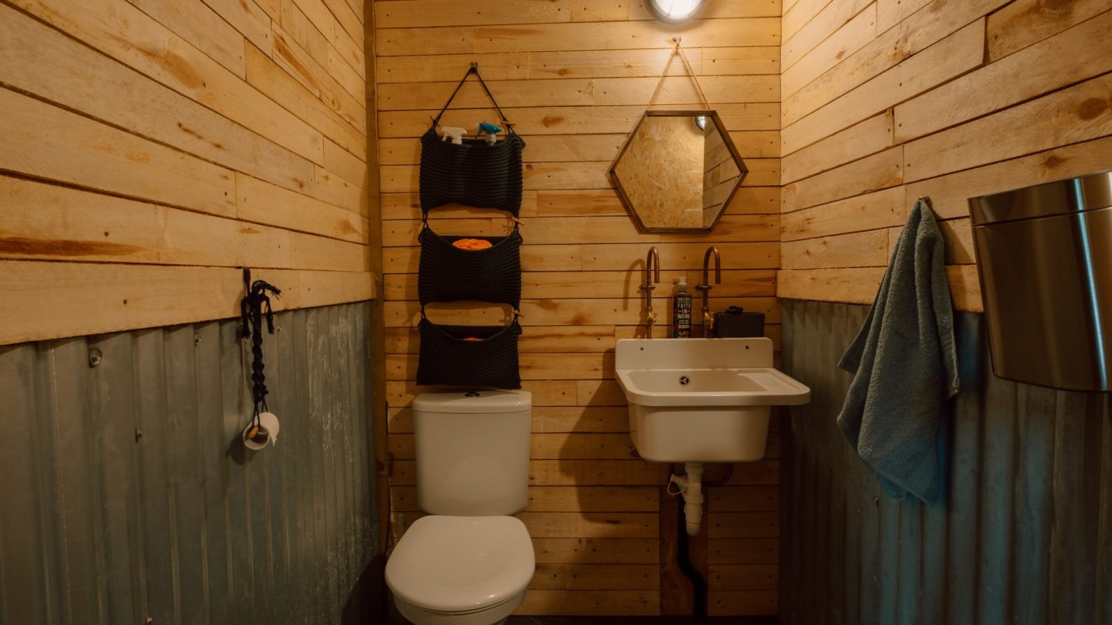 A small rustic bathroom at Y Caban at CampUs with a wooden wall, white toilet, wall-mounted sink, and hexagonal mirror.