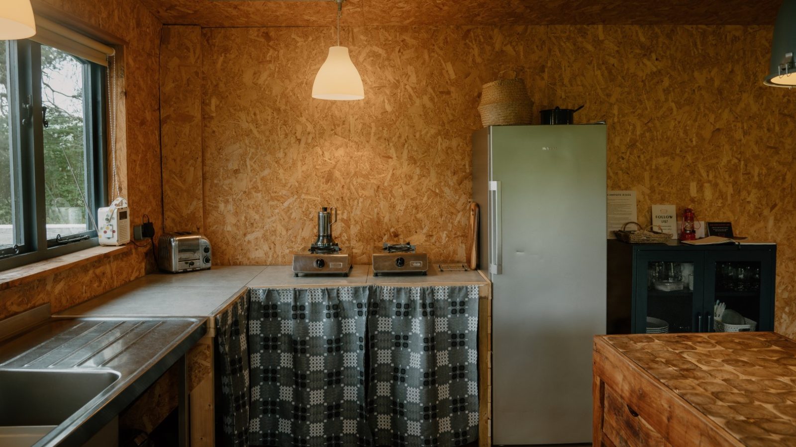 Rustic kitchen at Y Caban with wooden walls, a fridge, toaster, and two-burner stove on a counter with plaid curtains.