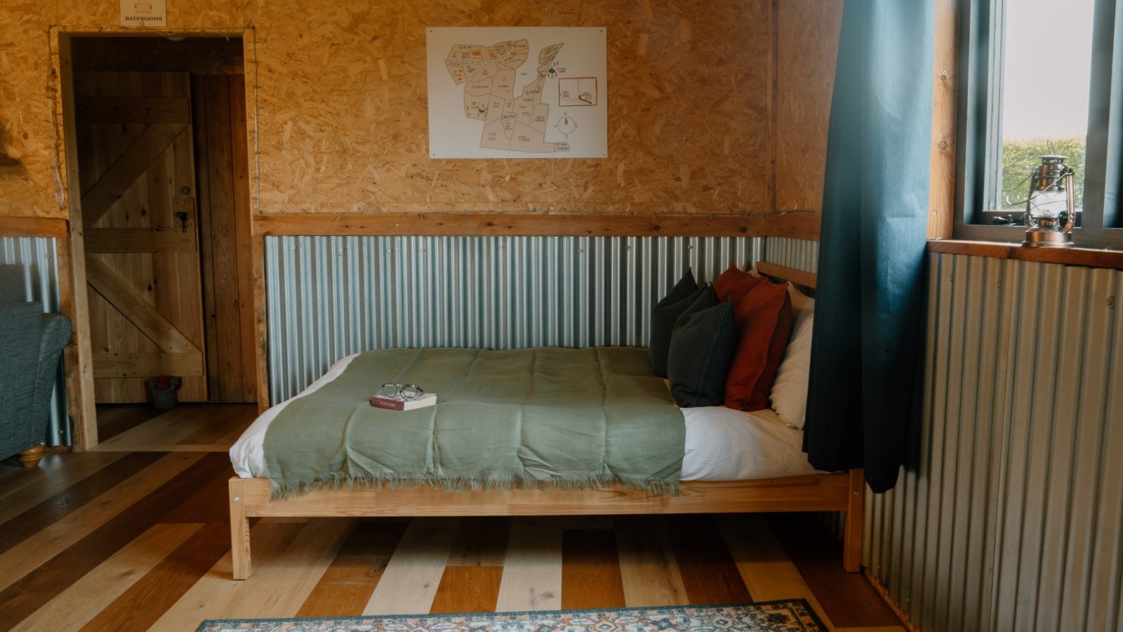 A cozy room at Y Caban with a wooden bed, green blanket, and a small framed map on wooden walls beside the curtained window.