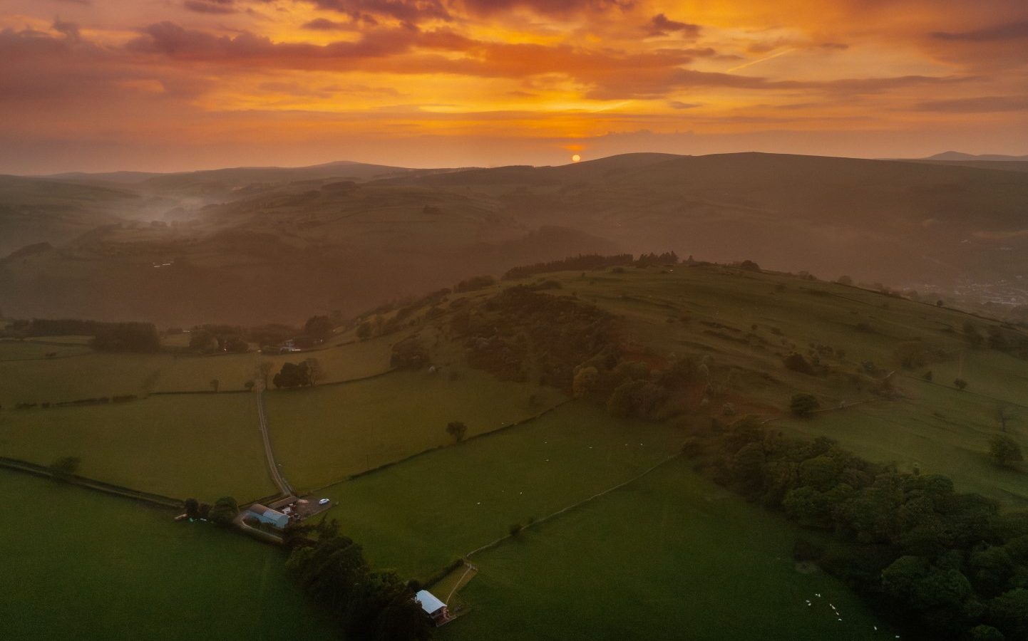 Aerial view of Y Caban at CampUs, nestled in a verdant landscape with rolling hills and a mesmerizing orange sunset sky.