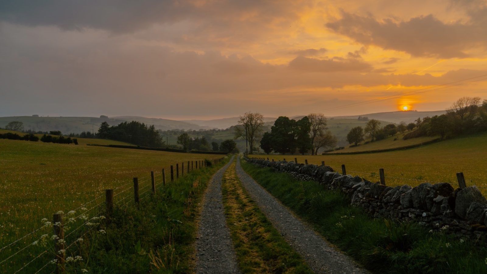 A dirt path at Y Caban leads through fields at sunset, flanked by stone walls under a cloudy sky with an orange glow.