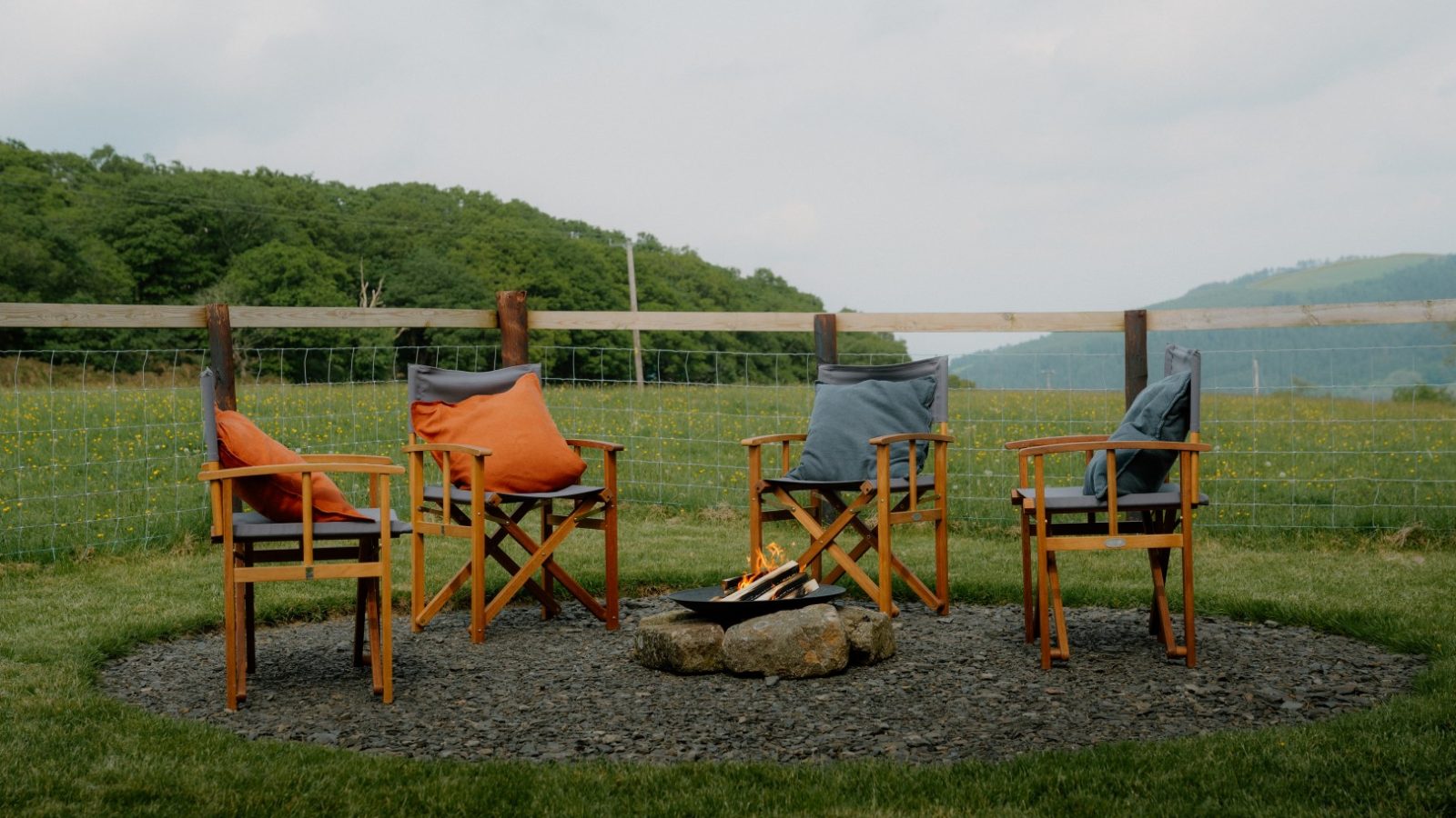 Four wooden chairs with orange and gray cushions encircle the fire pit at Y Caban at CampUs, nestled in a rural landscape.