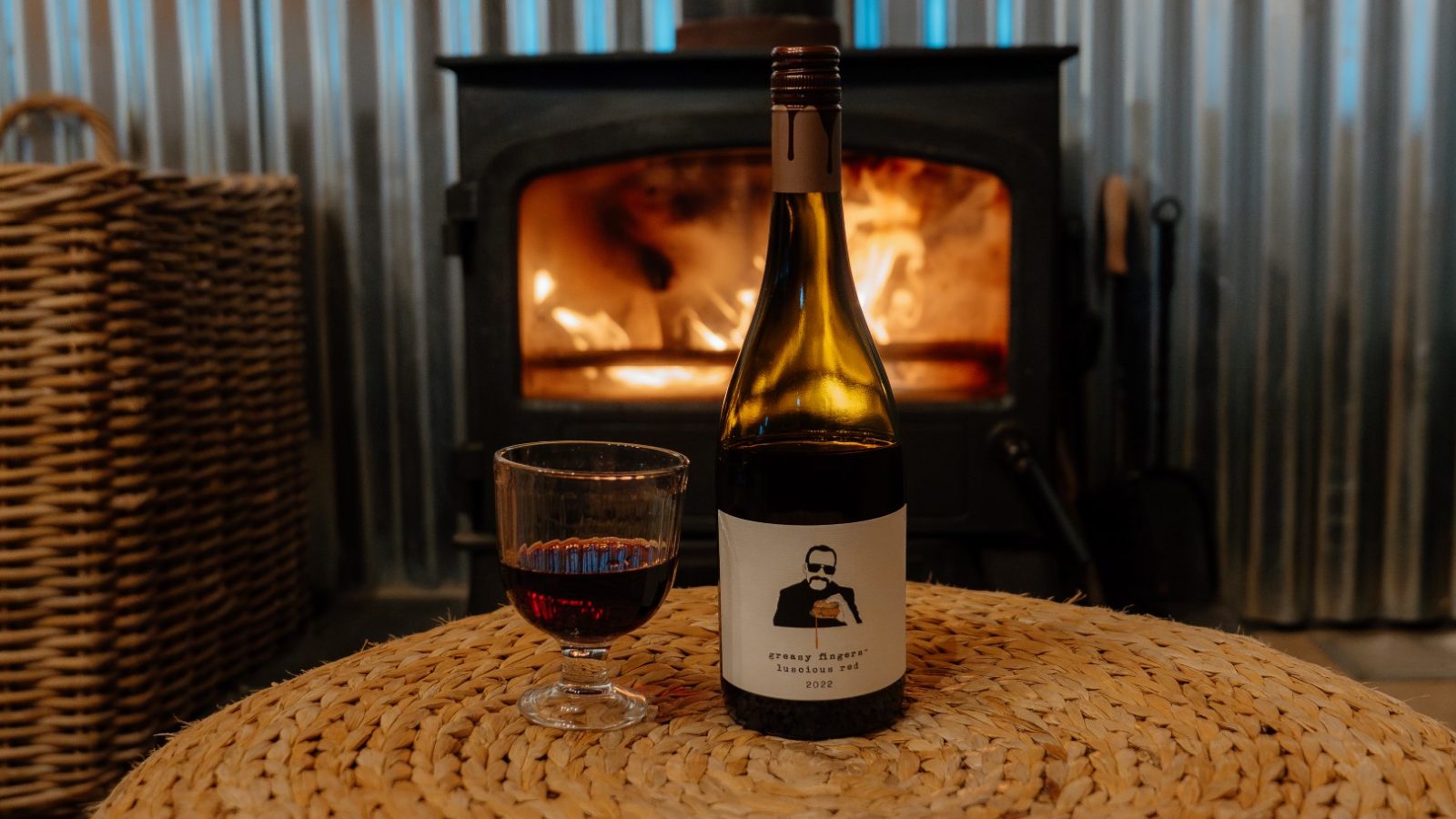 A bottle of wine and filled glass rest on a woven mat in front of the warm wood stove at Y Caban at CampUs.