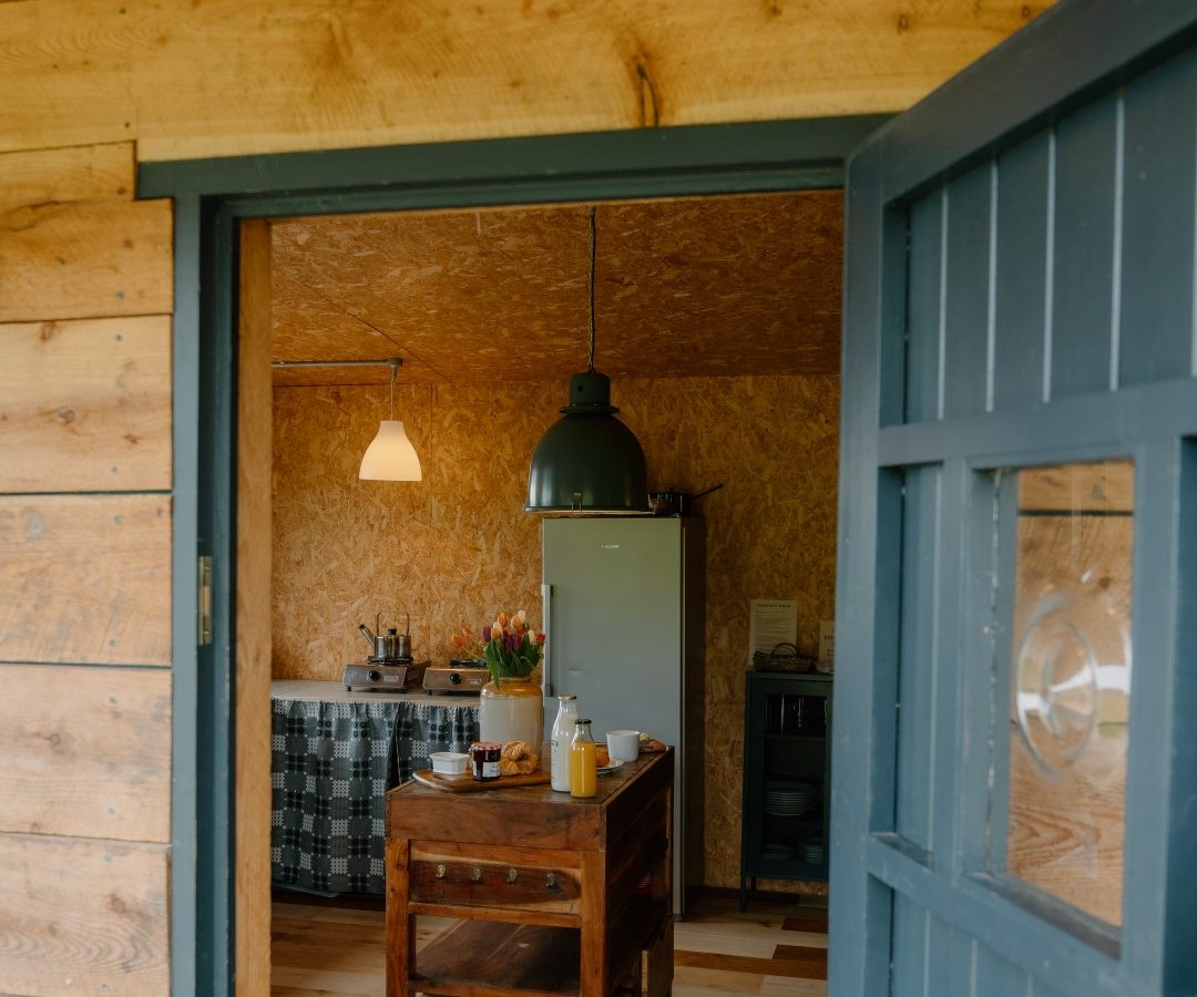 Y Caban at CampUs: A wooden cabin entrance with door ajar, cozy interior, kitchenette, fridge, and breakfast on the table.