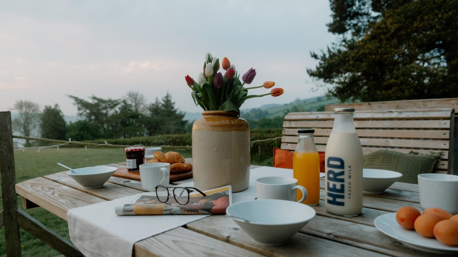 Outdoor breakfast at Y Caban at CampUs: bowls, plates, juice, croissants, and tulips with a scenic view.